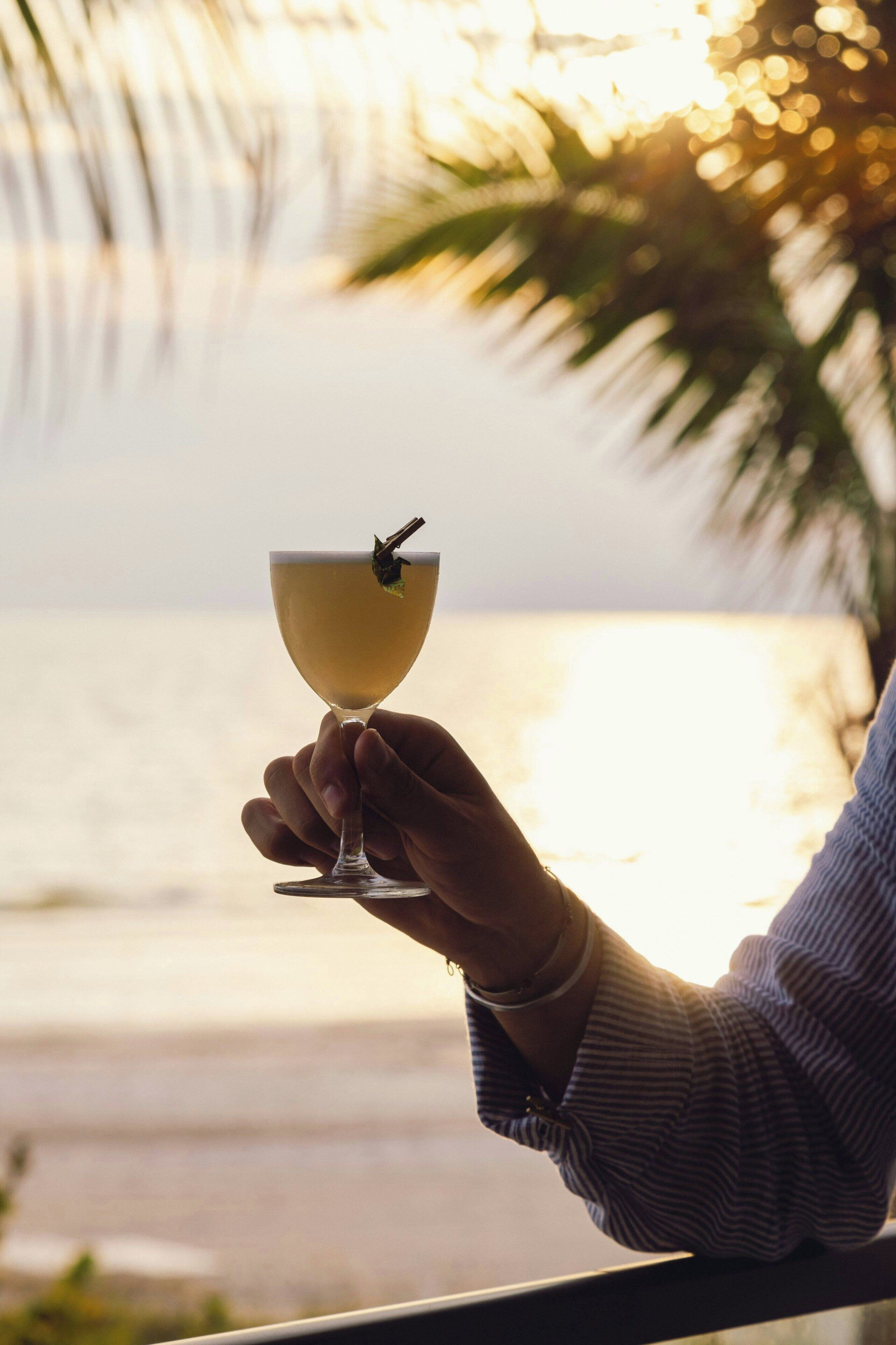 En hand håller i ett cocktailglas med en dekoration, och i bakgrunden syns en strand med palmer.