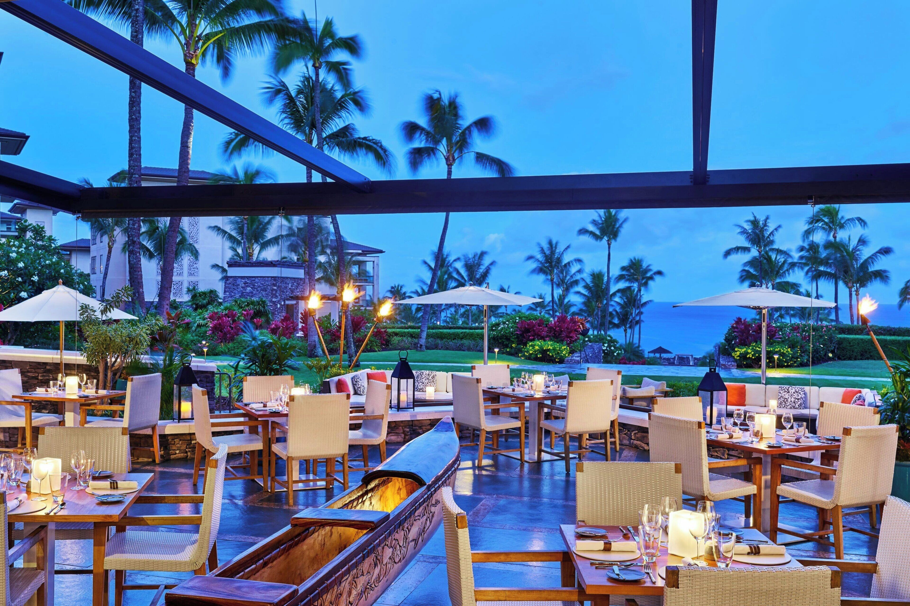 An outdoor dining area with tables, chairs, and umbrellas, overlooking a landscaped garden and palm trees.