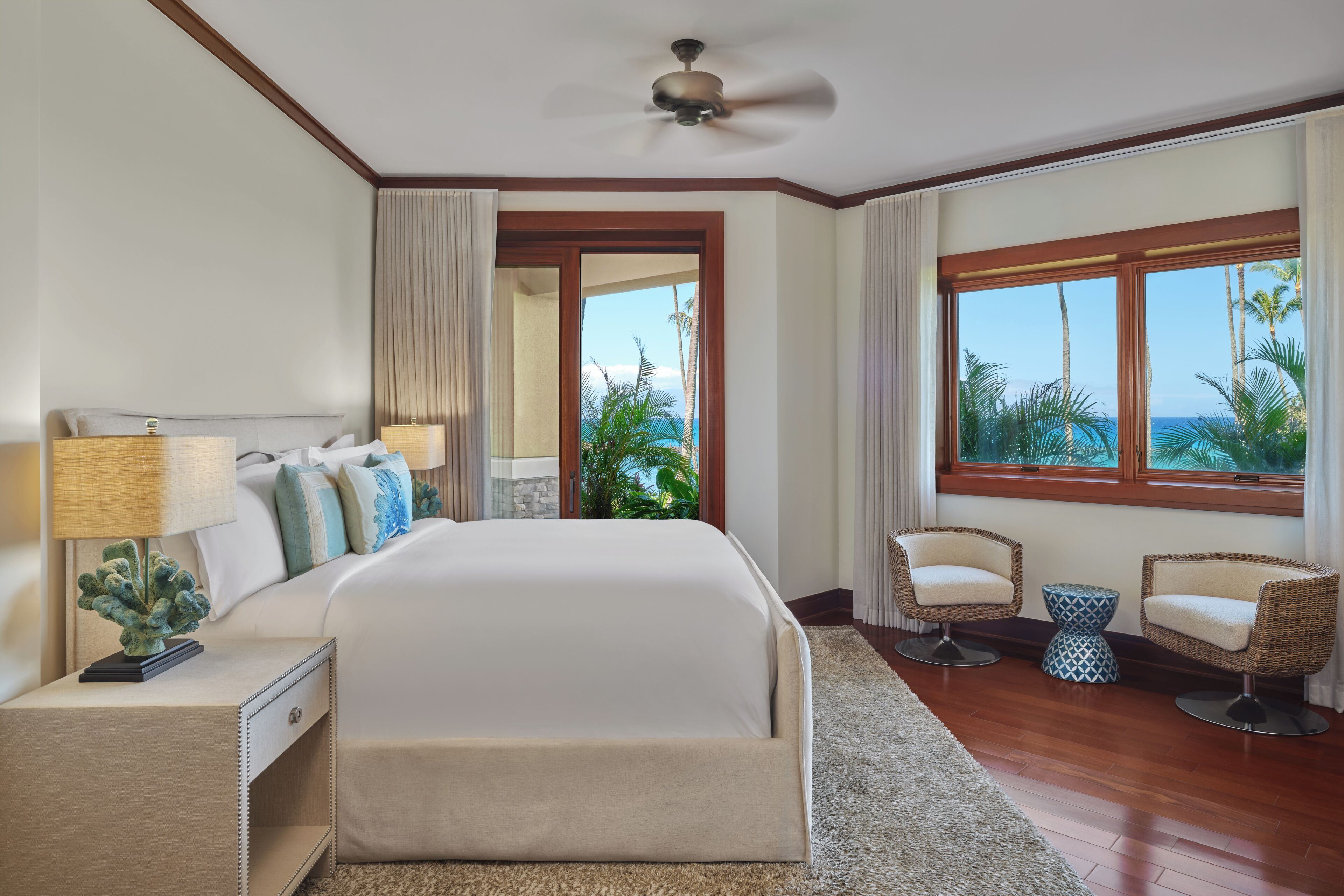 A bedroom with a bed, nightstands, a lamp, two chairs, a window with a view, and a ceiling fan.