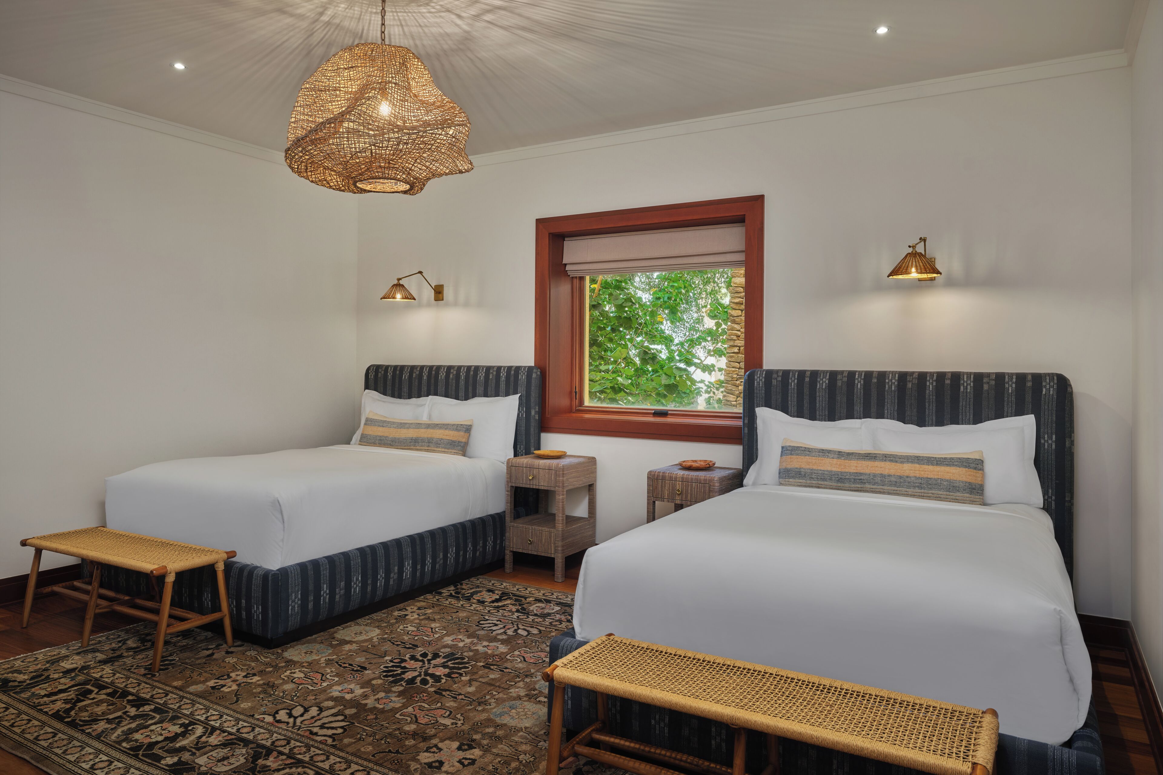 A hotel room with two beds, a woven ceiling light, a window with a view, and a patterned rug.