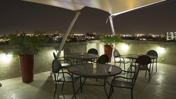 An outdoor patio with round tables and chairs, potted plants, and a city view at night. - Hotel Innova Chipichape (Cali)