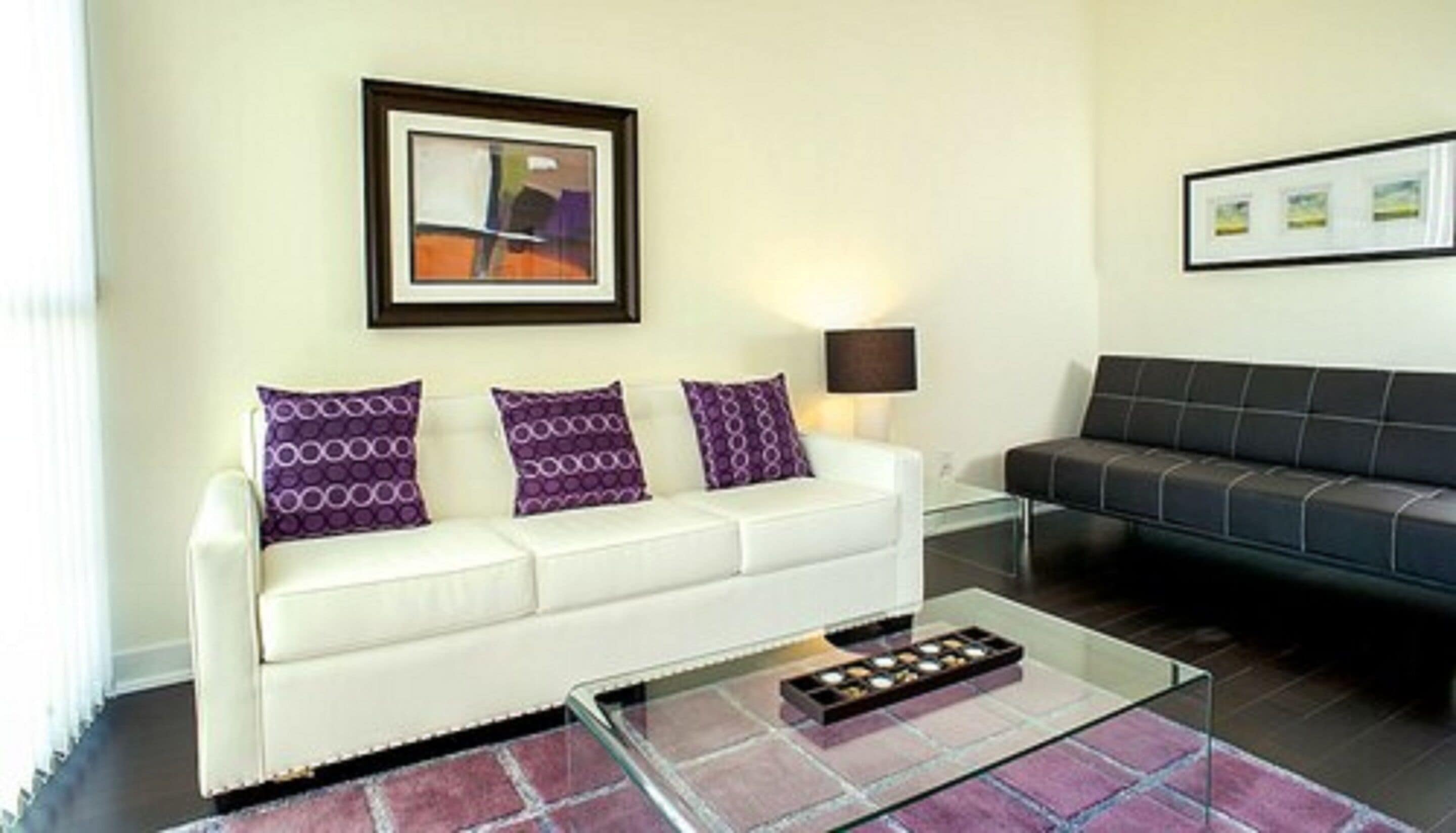 A modern living room with a white sofa, a black sofa, and a glass coffee table. There are three purple pillows on the white sofa.