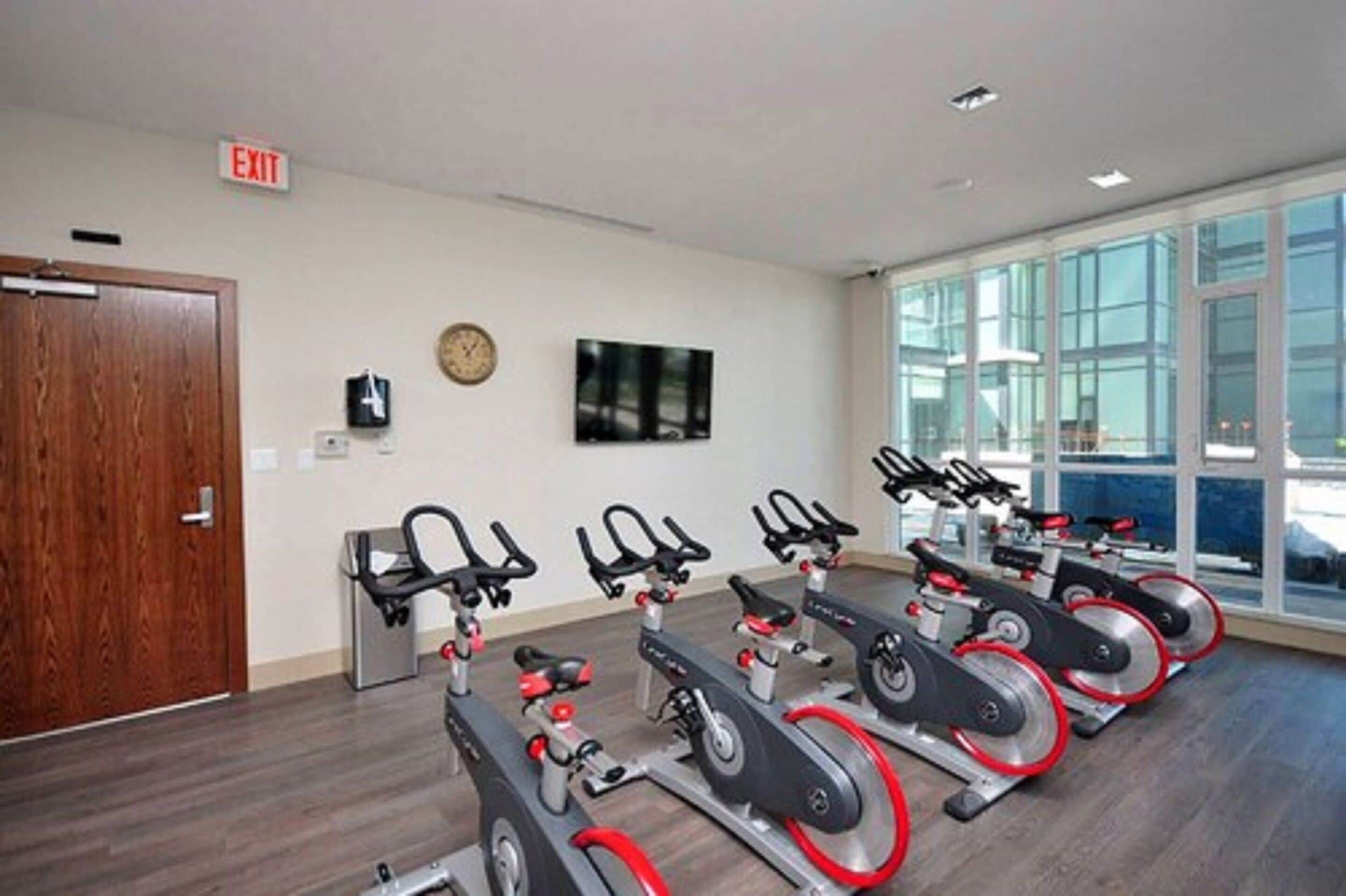 A gym with four stationary bikes, a clock, and an exit sign.