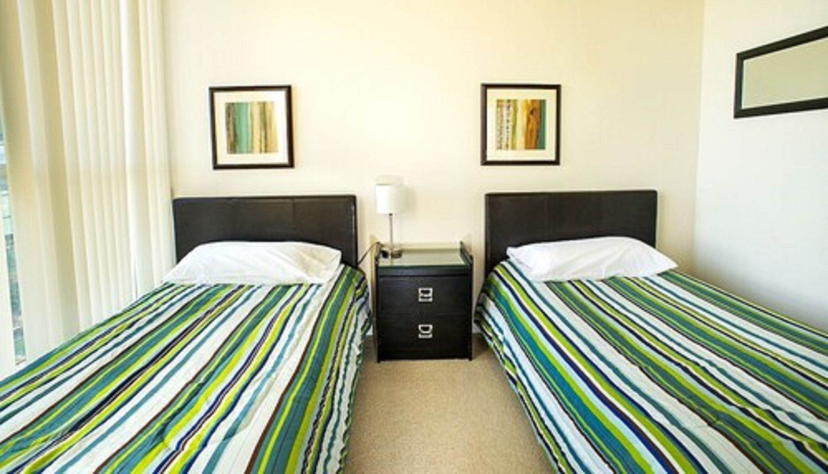 Une chambre d’hôtel avec deux lits simples, chacun doté de draps à rayures, une table de chevet avec une lampe et une petite commode.