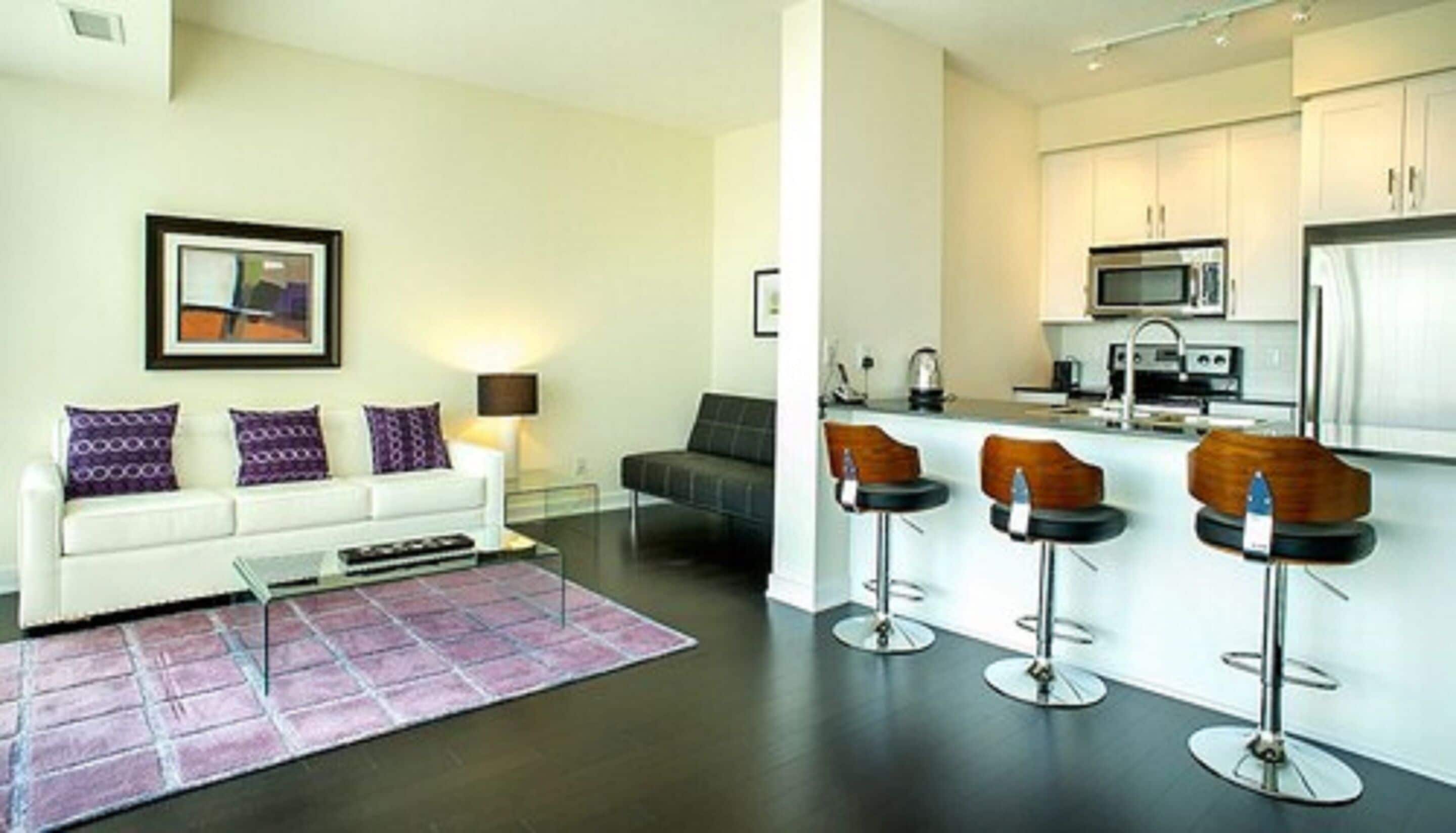 A modern living room with a white sofa, purple cushions, and a coffee table. There is a kitchen area with a microwave, stove, and bar stools.