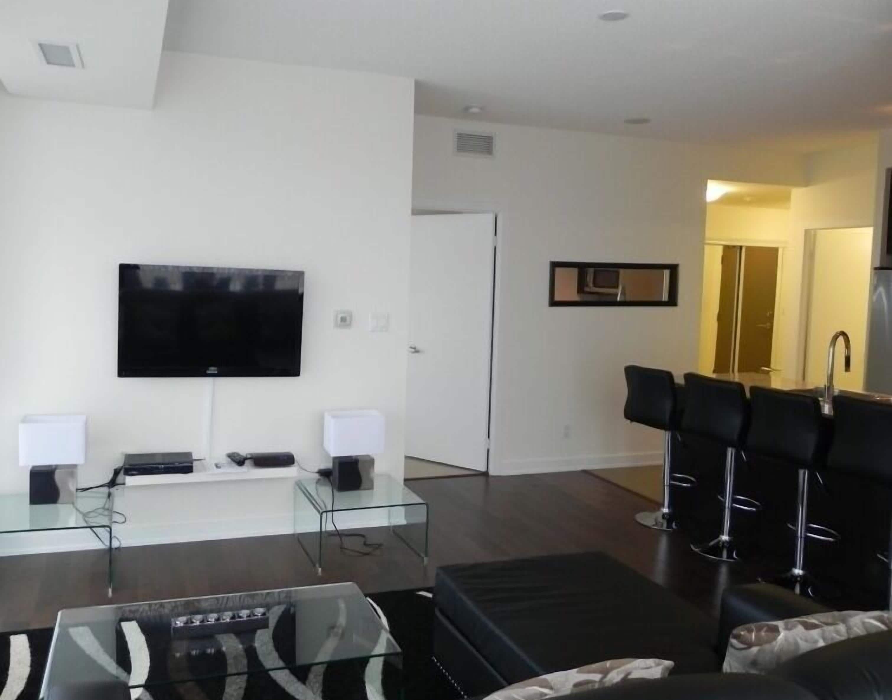 A modern living room with a flat-screen TV, a glass coffee table, and a seating area with black chairs.