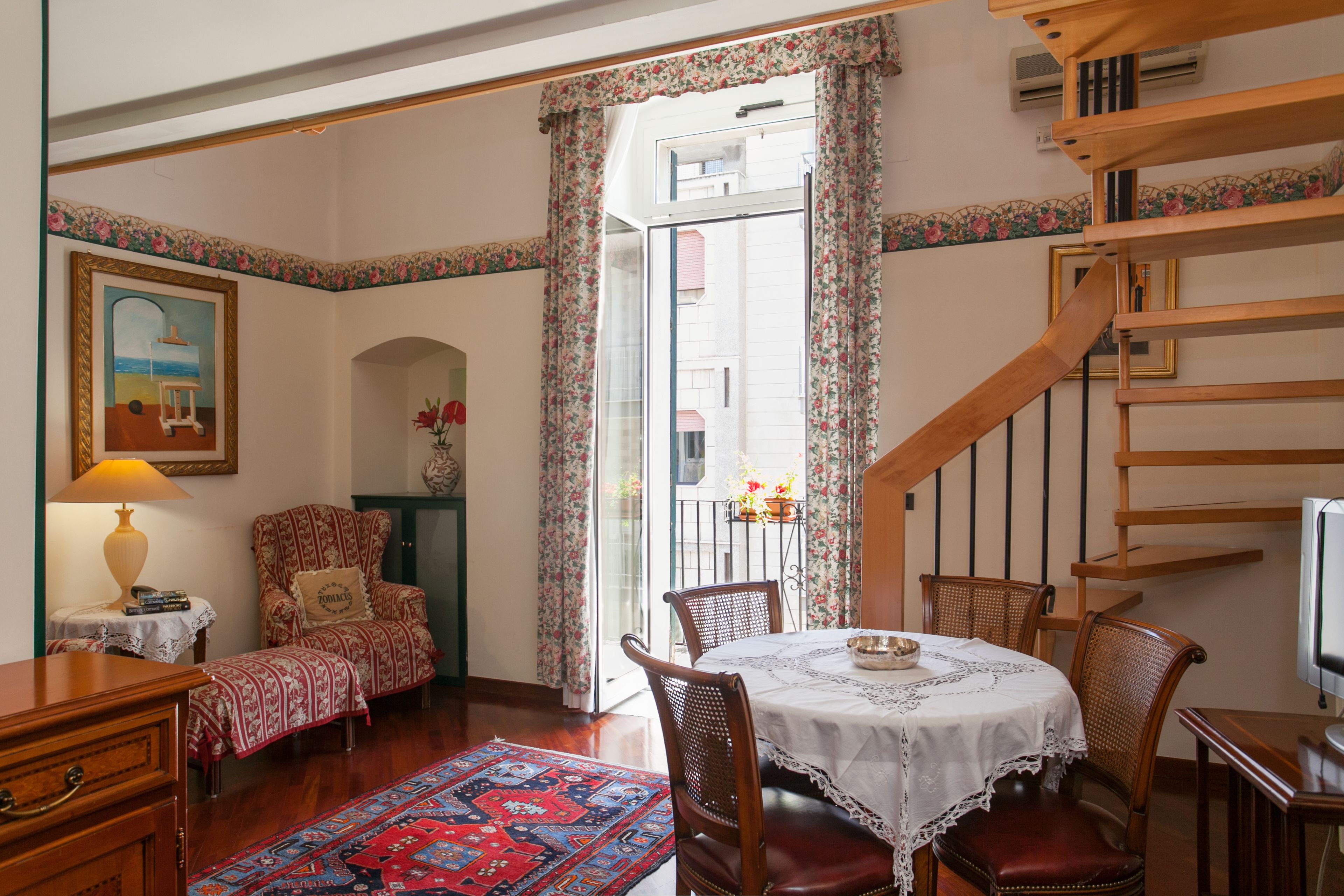 A dining area with a table set for two, surrounded by wooden chairs, a patterned rug, and a painting on the wall.