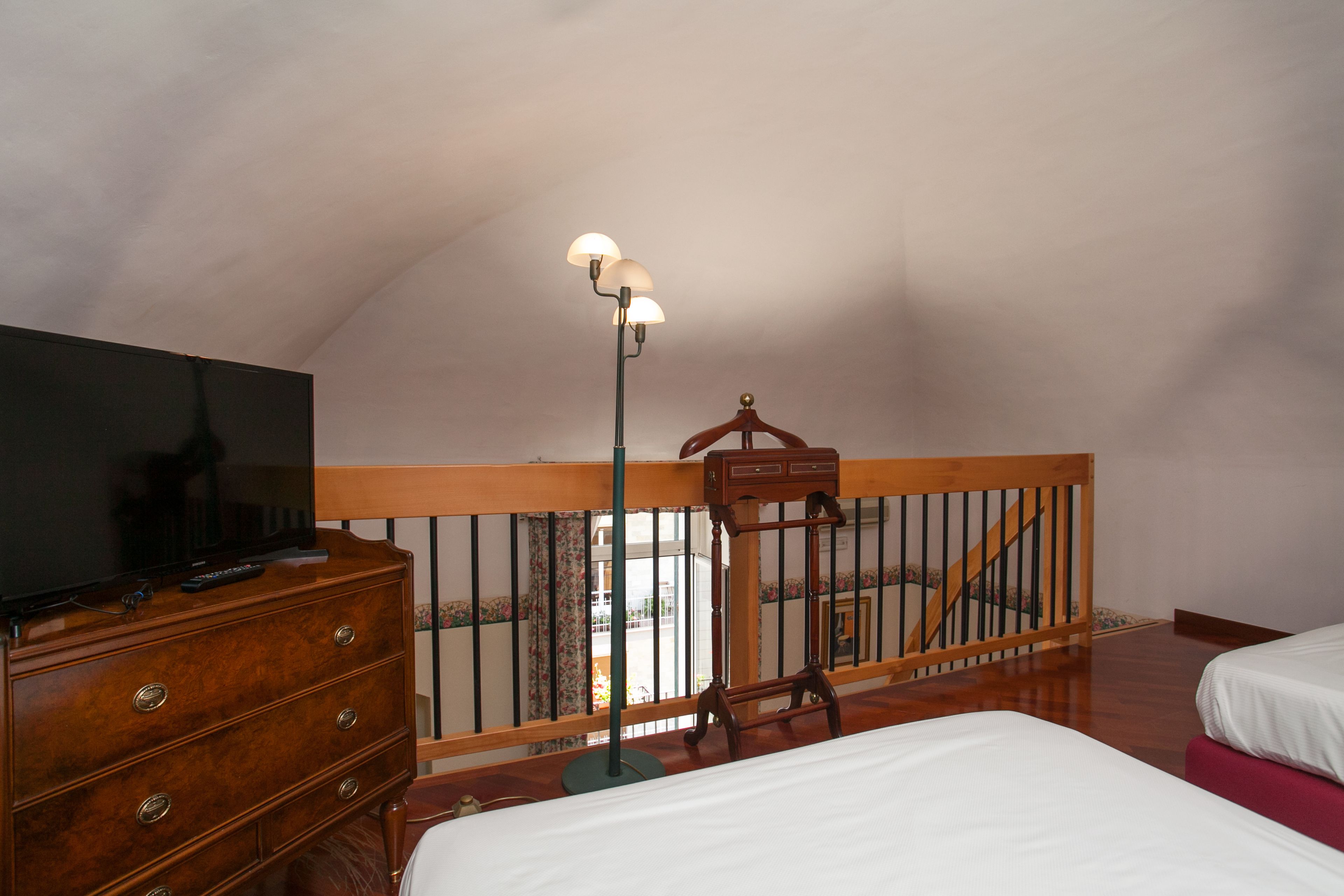 Quarto com cômoda de madeira, uma cama, mesa de cabeceira, abajur e guarda-corpo de varanda.