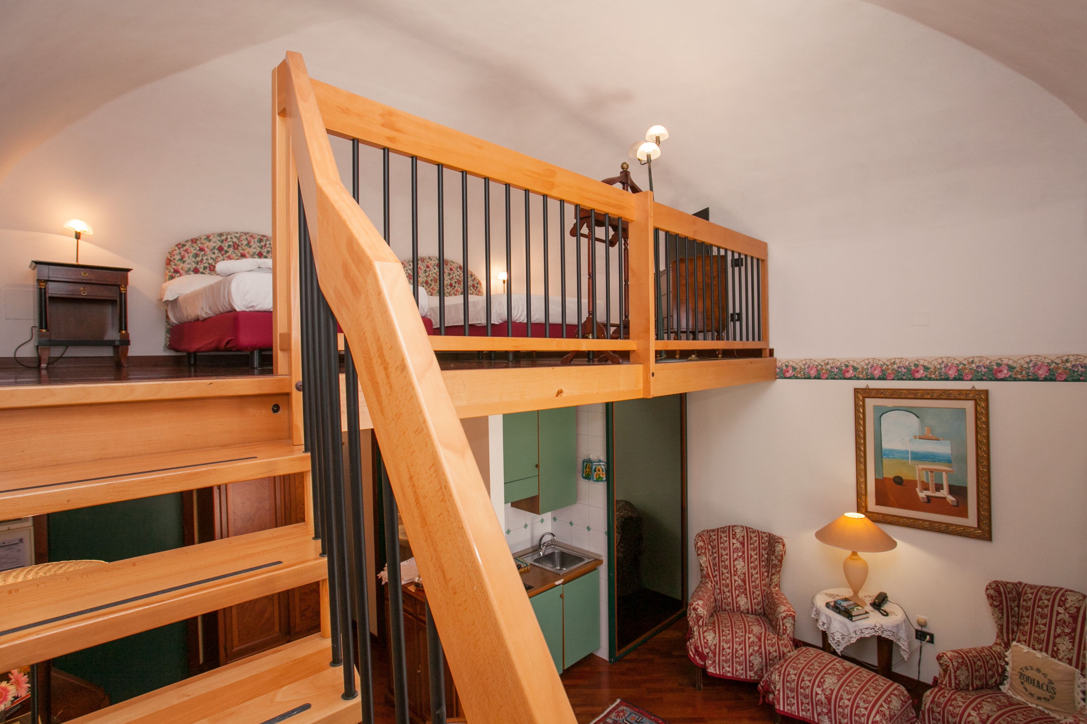 Quarto no estilo loft com escada de madeira, cama com roupa de cama floral, pequeno espaço de cozinha e cadeiras estampadas.