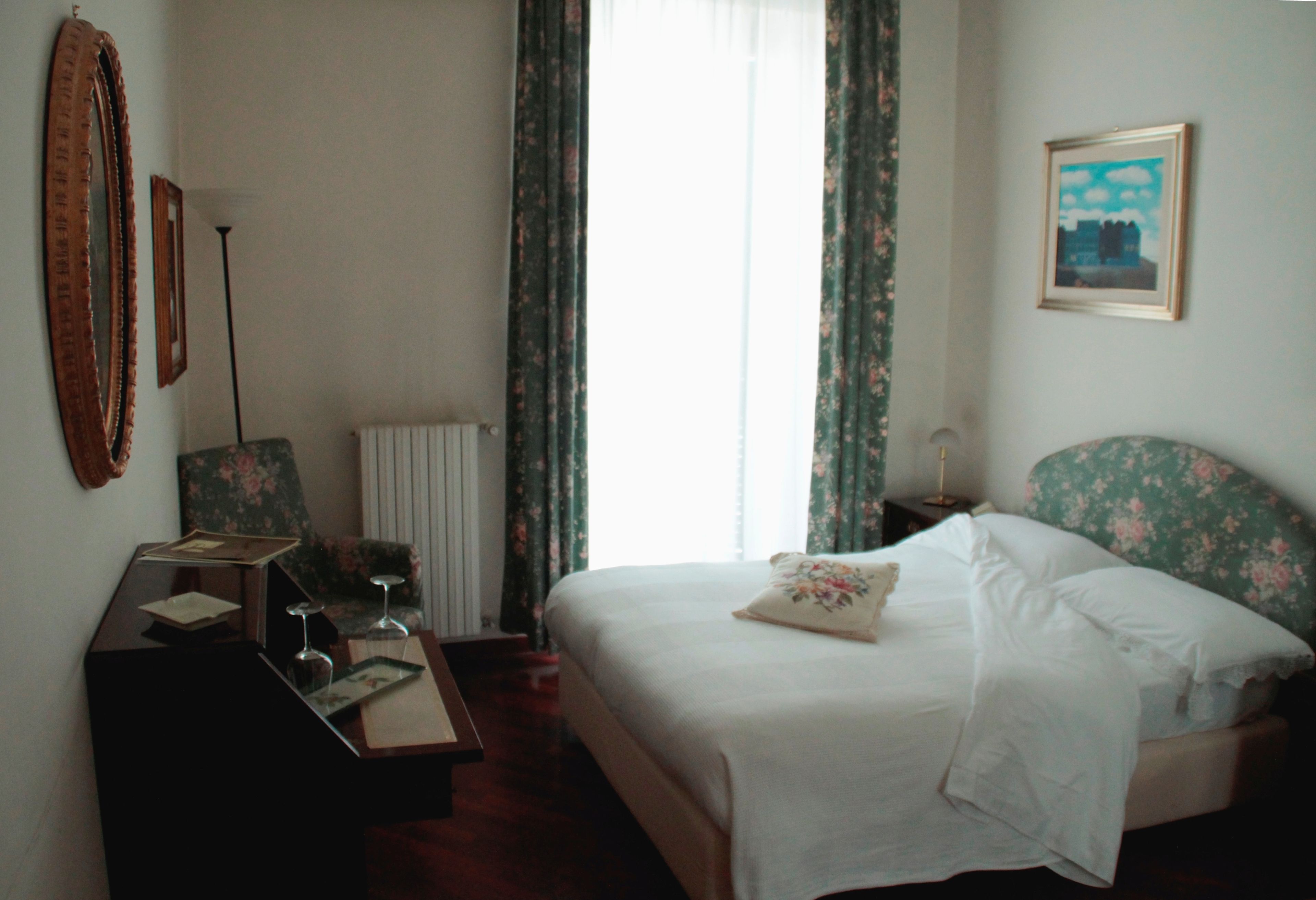 A hotel room with a bed, a desk, a chair, a radiator, and a window with curtains.