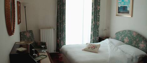 A hotel room with a bed, a desk, a chair, a radiator, and a window with curtains.