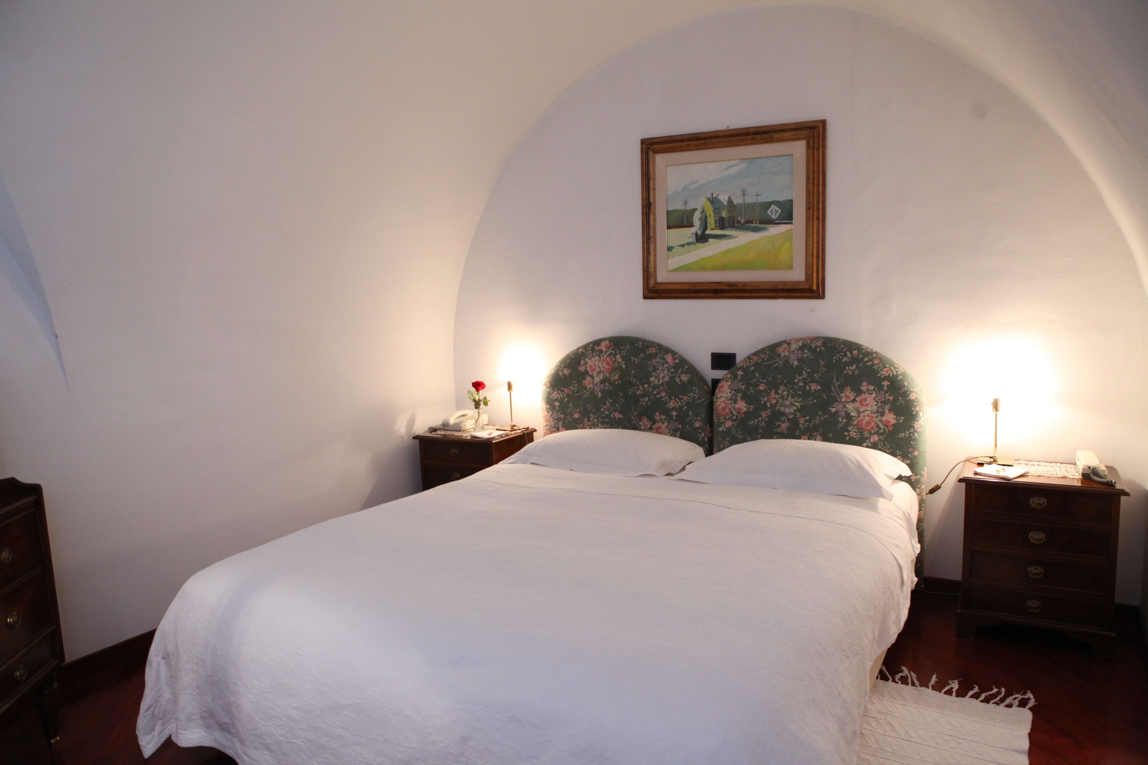 Una cama bien hecha con edredón blanco en una habitación con techo curvo y papel tapiz floral. Hay dos mesitas de noche con lámparas y un cuadro en la pared sobre la cama.
