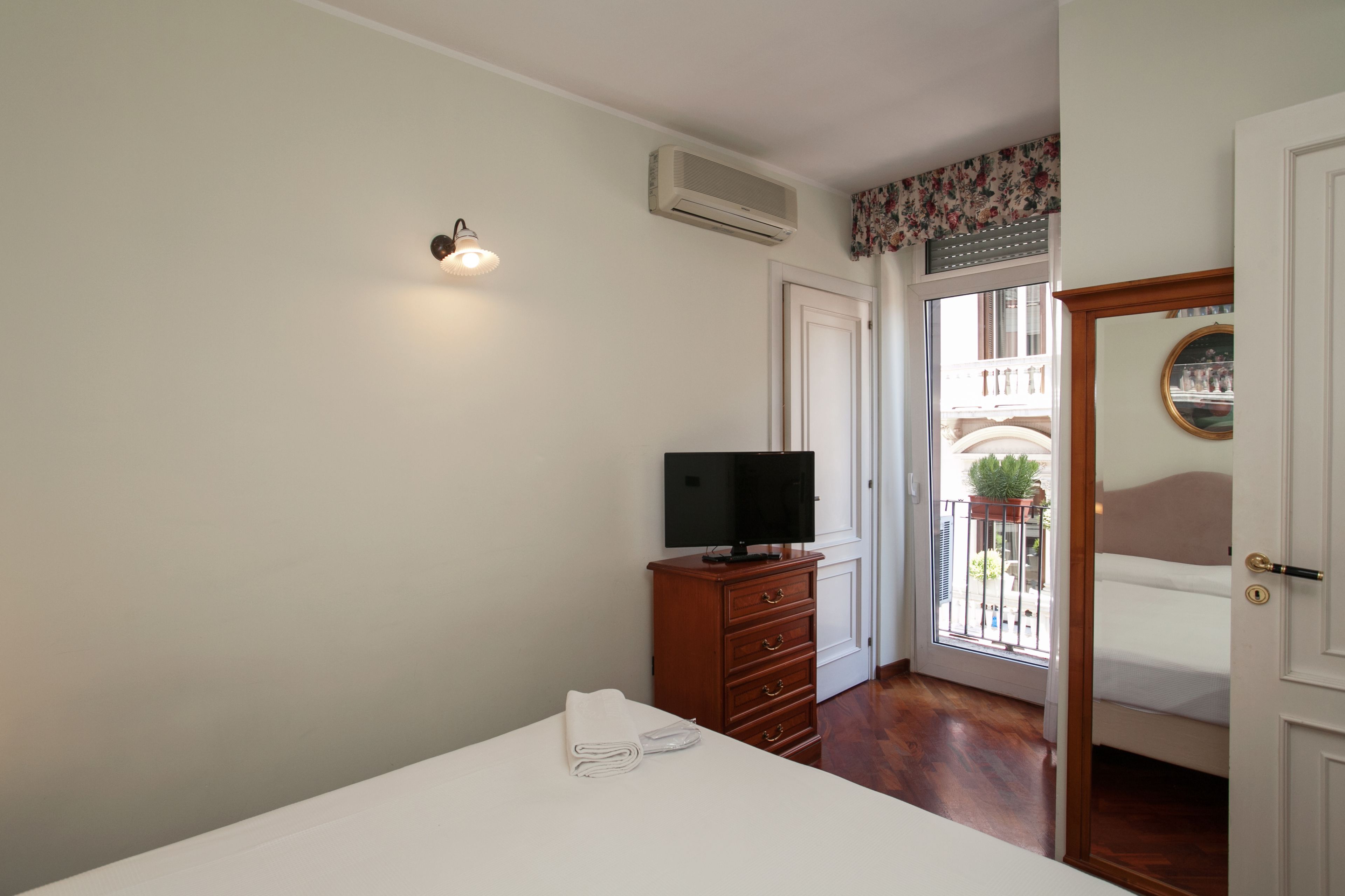 A bedroom with a bed, a TV on a wooden dresser, a door leading to a balcony, and a wall-mounted air conditioner.