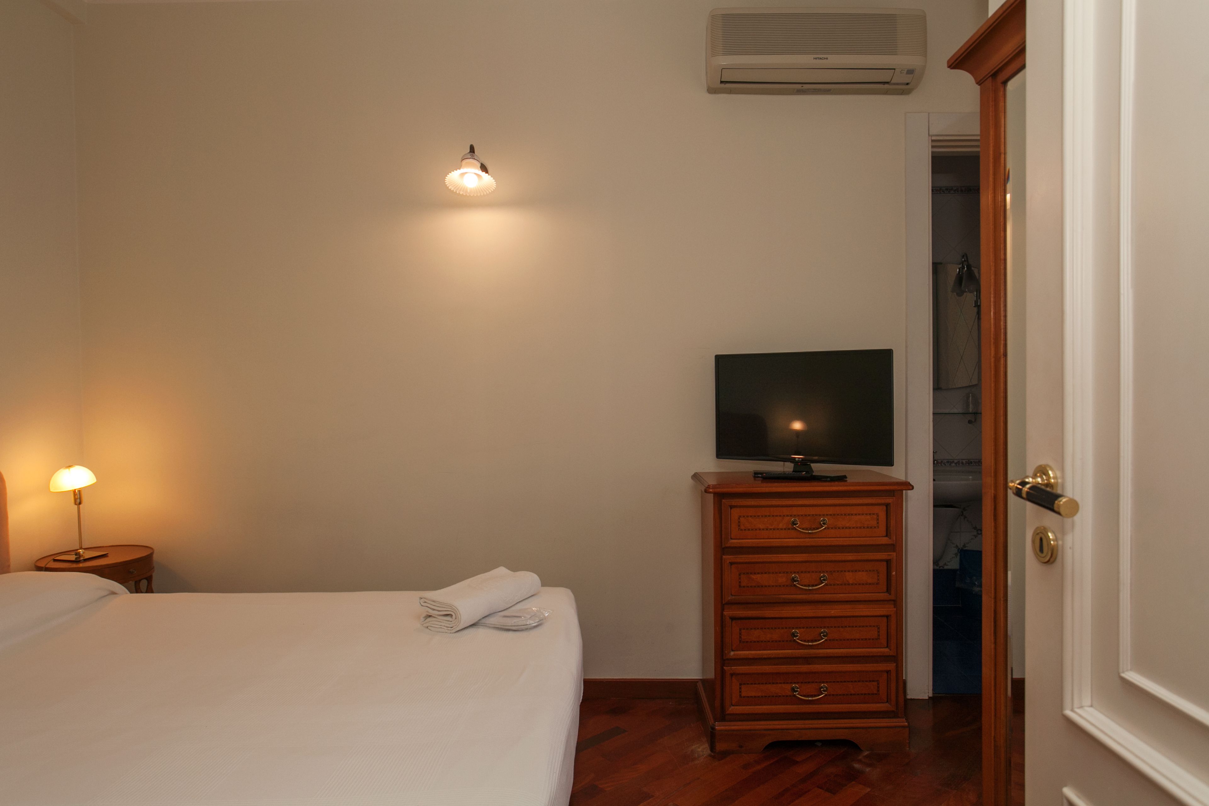 A bedroom with a bed, a TV on a wooden dresser, a wall-mounted air conditioner, and a door to another room.
