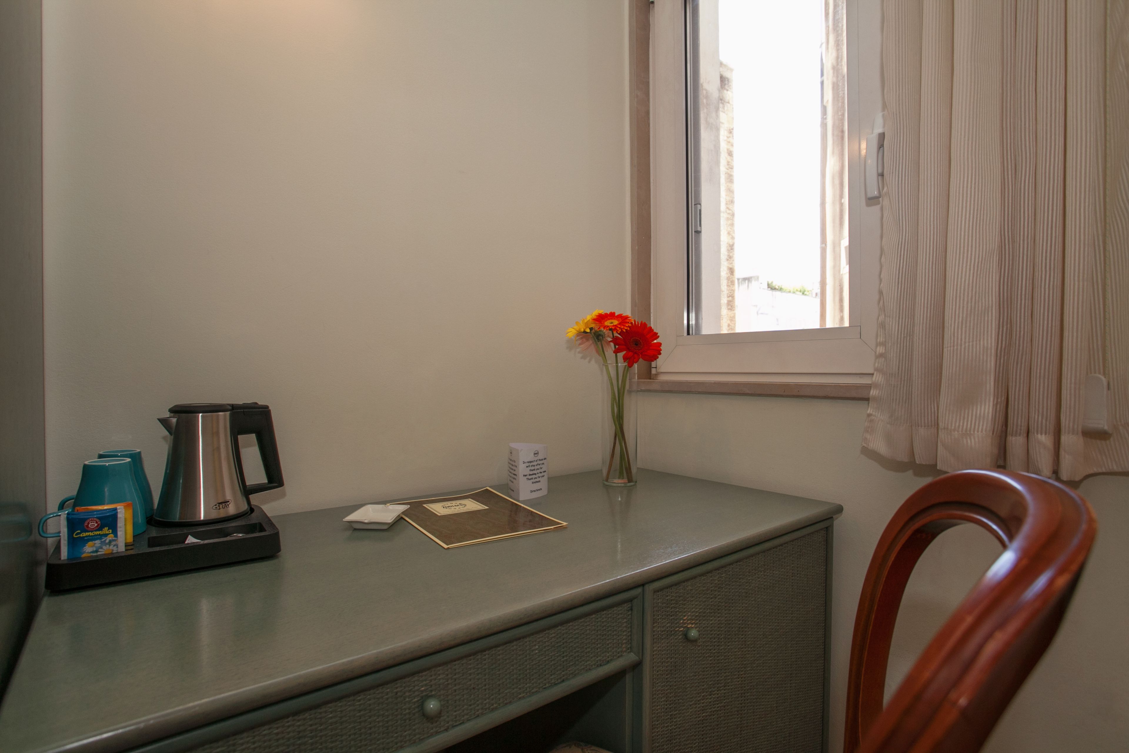 A small desk with a kettle, cups, a vase with flowers, and a window with curtains.