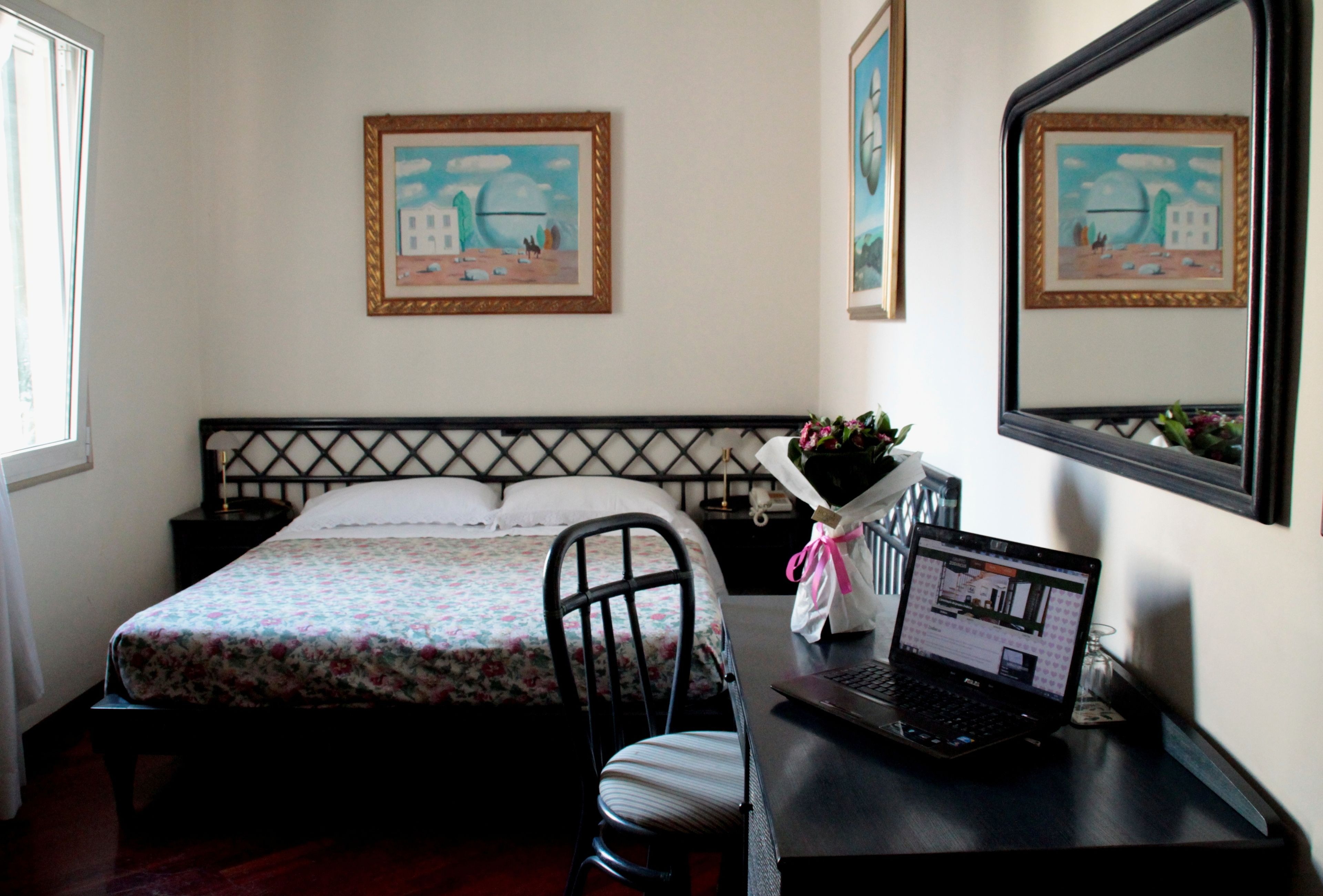 Een slaapkamer met een bed, een bureau met een laptop, een stoel en twee kunstwerken in lijsten aan de muur.
