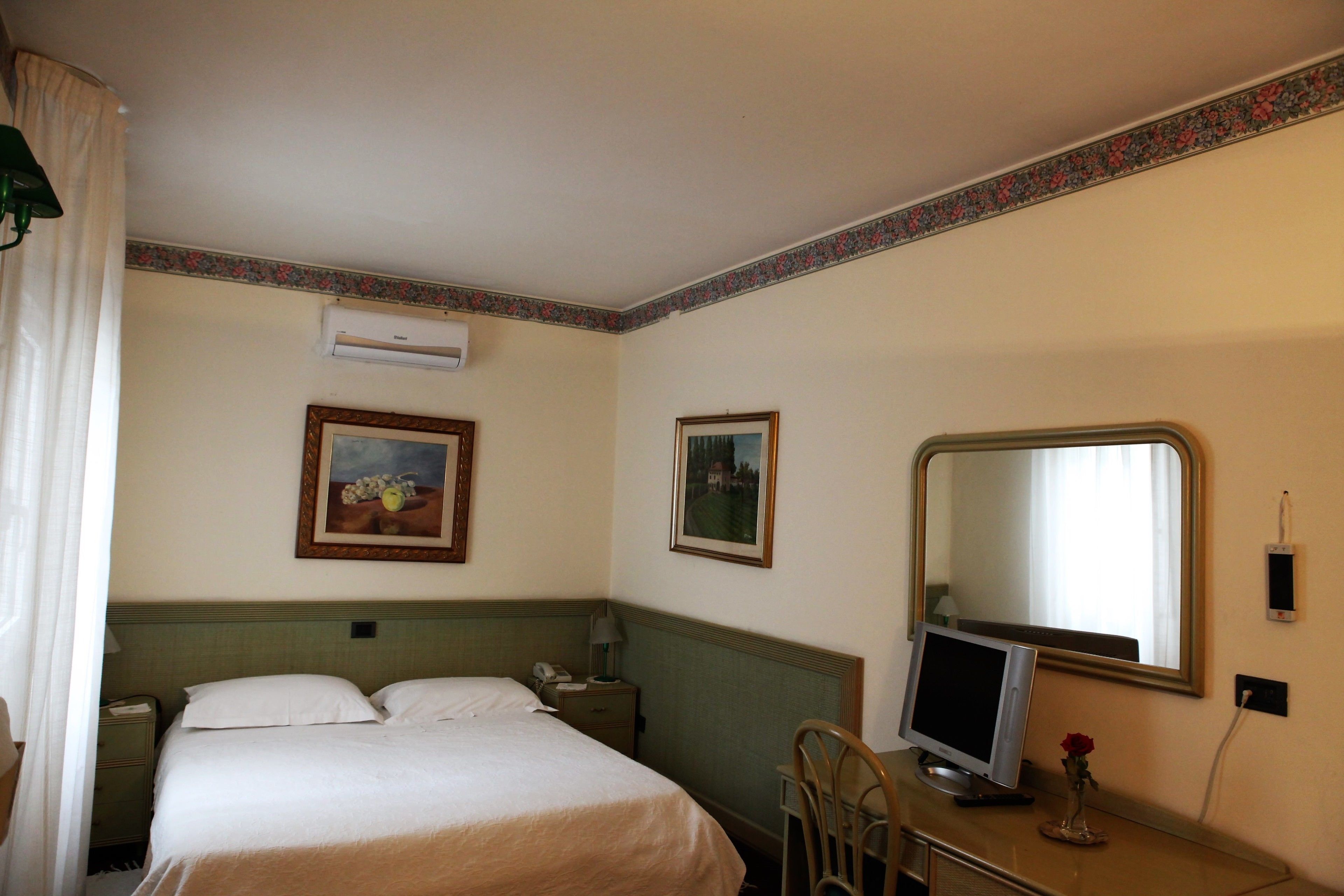 A hotel room with a bed, a desk with a computer, a telephone, and two framed pictures on the wall.