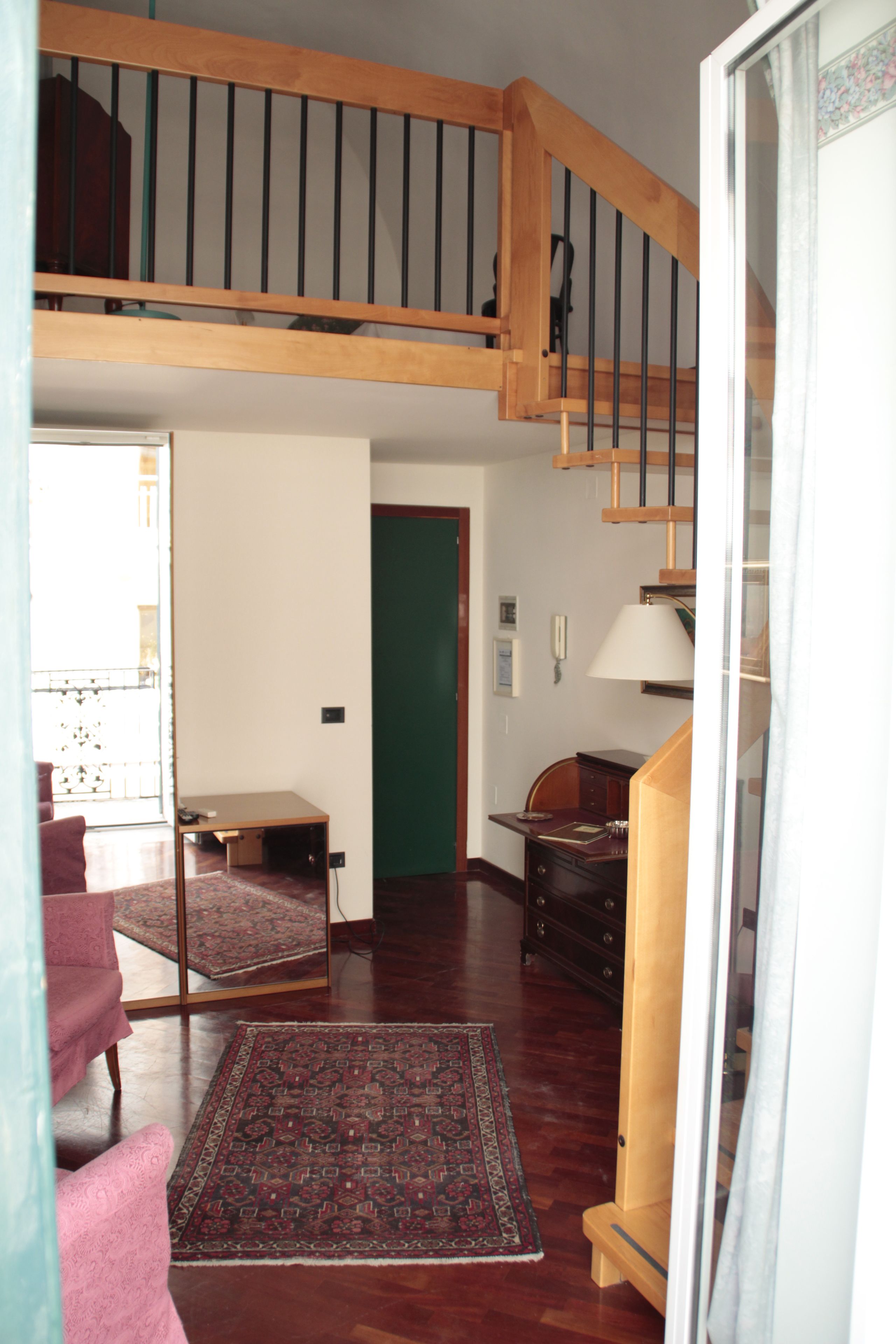 A spacious living room with a wooden staircase, a large rug, and a green door leading to another room.