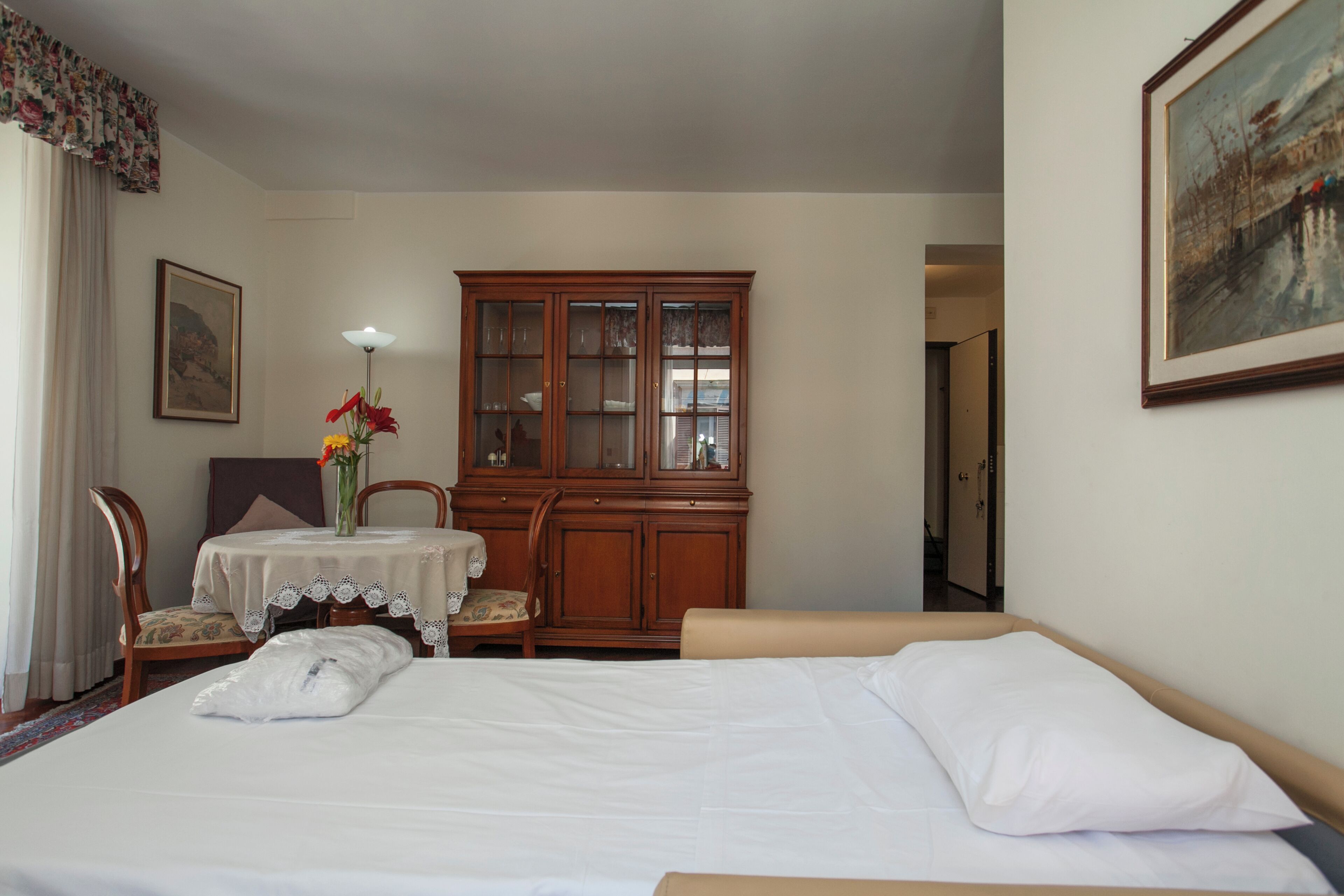 Una habitación de hotel con cama, un armario de madera, una mesa con un jarrón de flores y una silla.