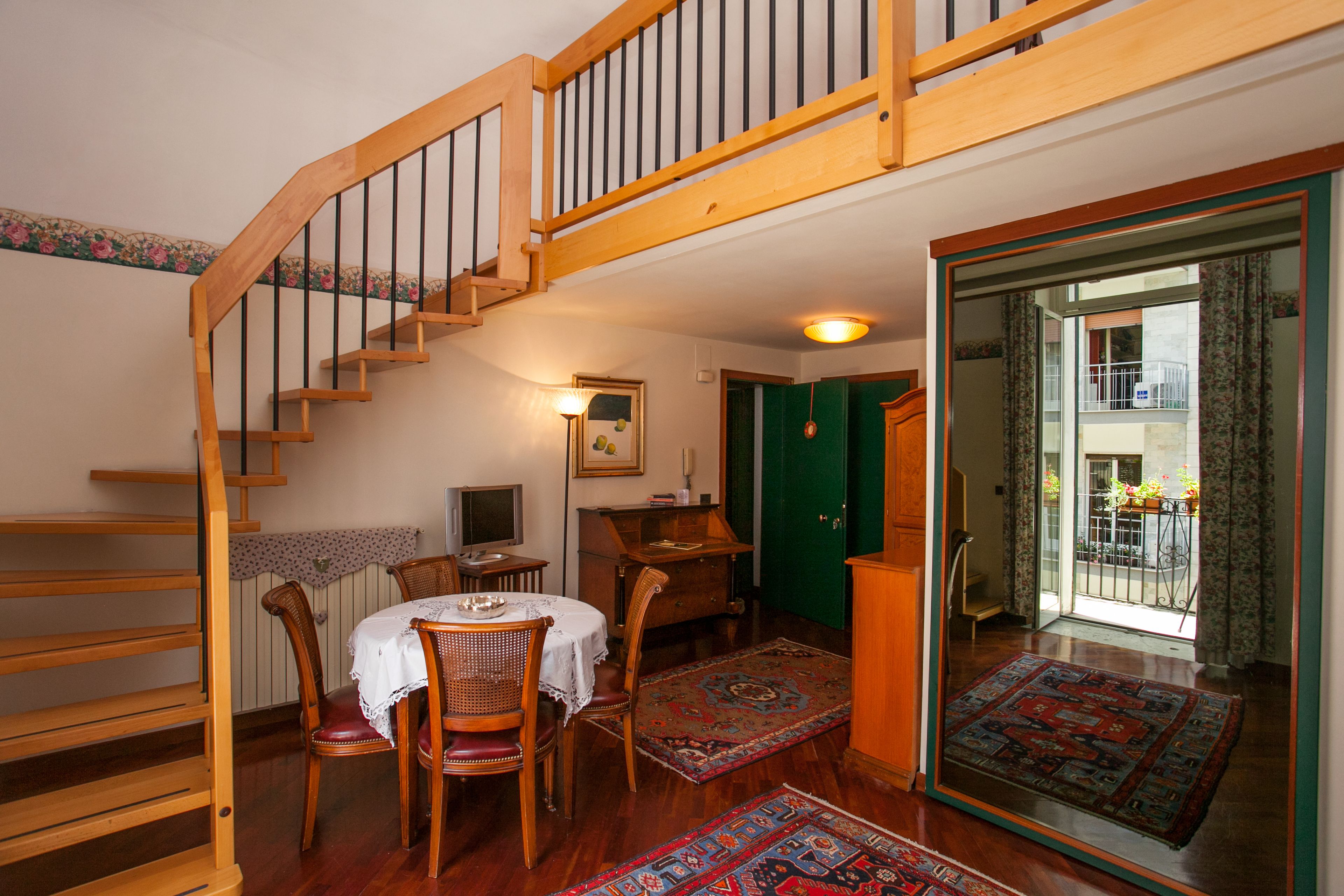 Una escalera de madera que lleva al segundo piso, un comedor con mesa y sillas, una puerta verde y vistas a un balcón con plantas.
