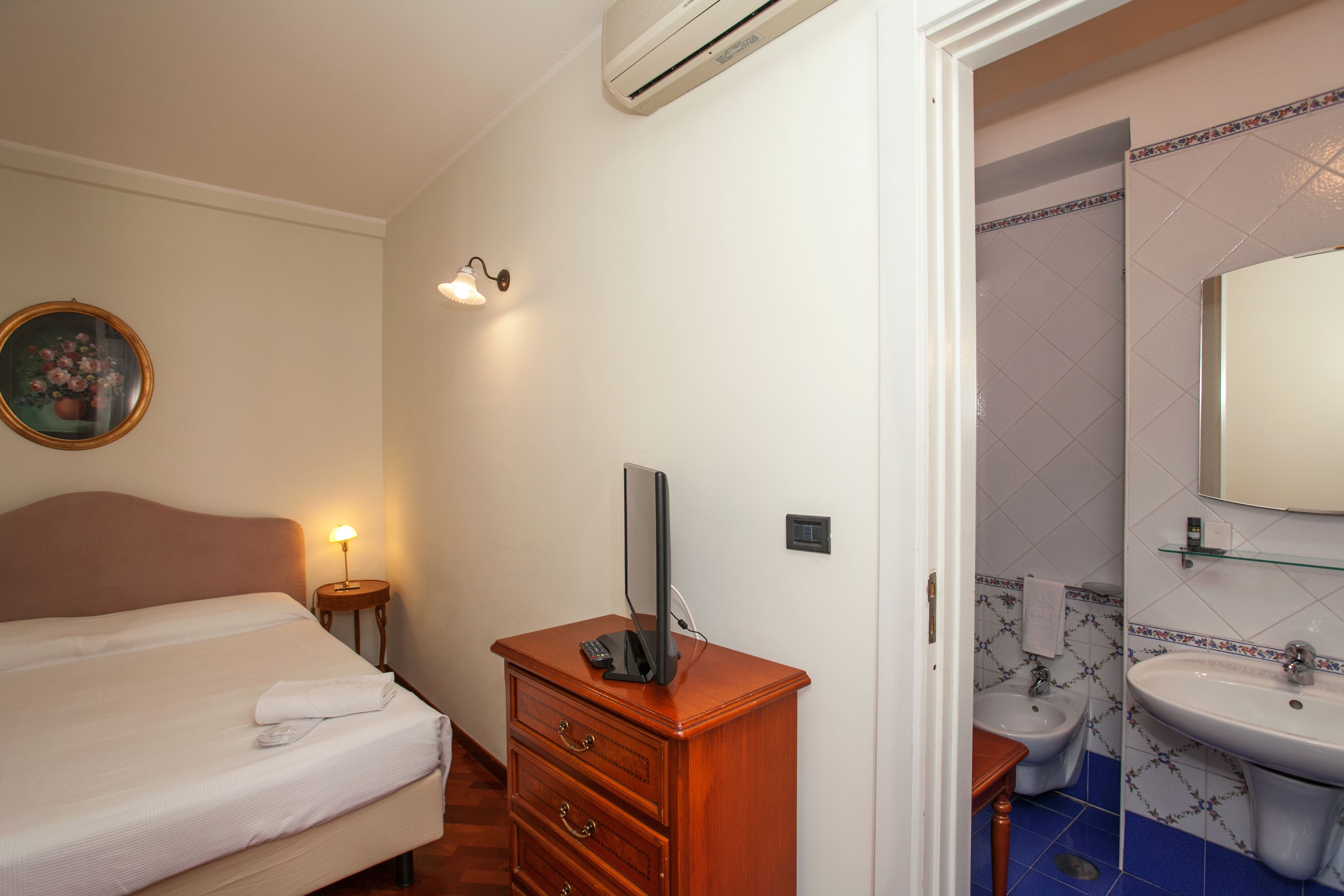A hotel room with a bed, a wooden dresser with a flat-screen TV, a bedside lamp, and a bathroom with a sink and bidet.