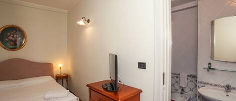 A hotel room with a bed, a wooden dresser with a flat-screen TV, a bedside lamp, and a bathroom with a sink and bidet.