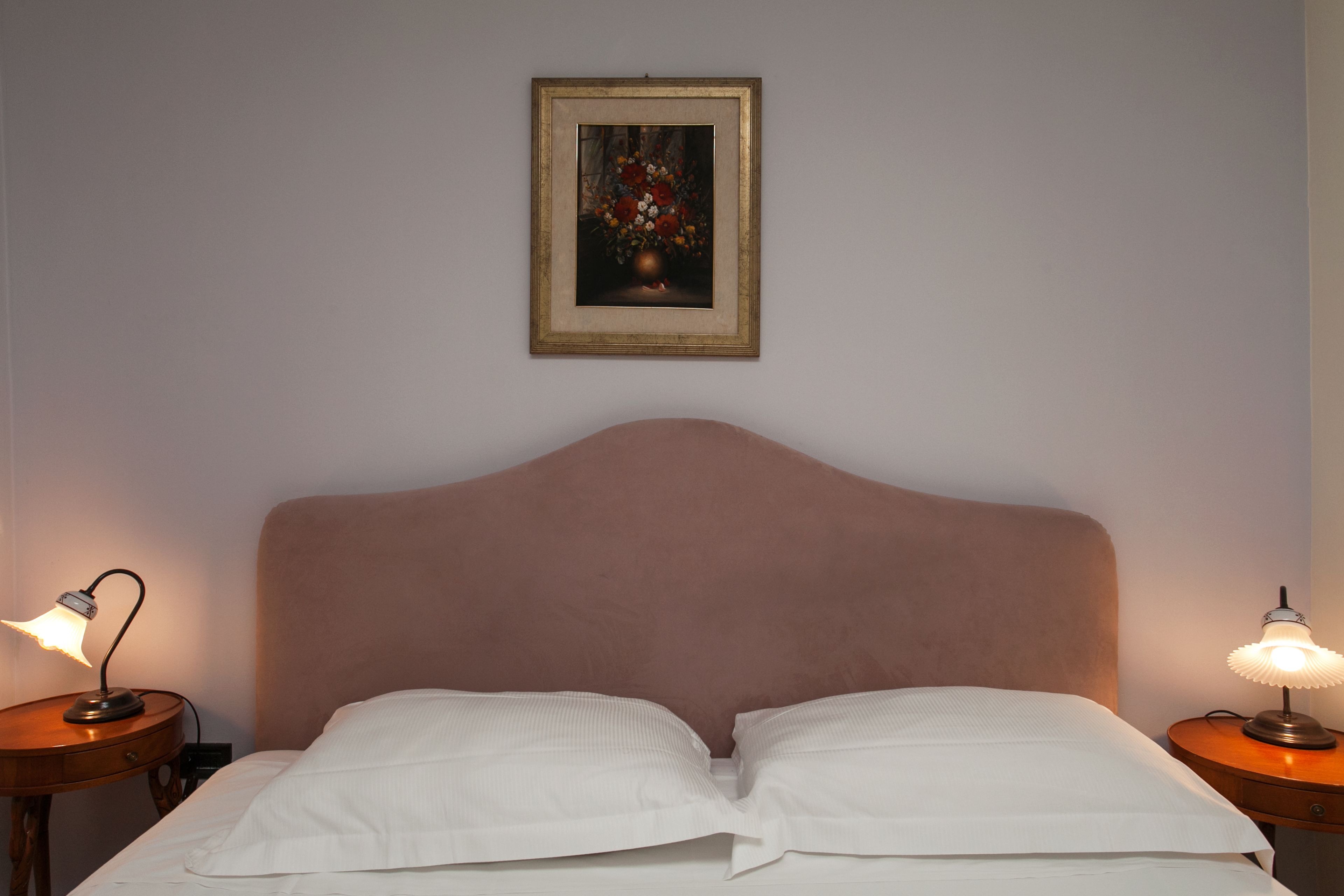 A neatly made bed with white linens, a headboard, two bedside lamps, and a framed floral artwork on the wall.