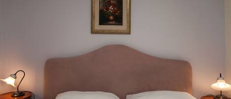 A neatly made bed with white linens, a headboard, two bedside lamps, and a framed floral artwork on the wall.