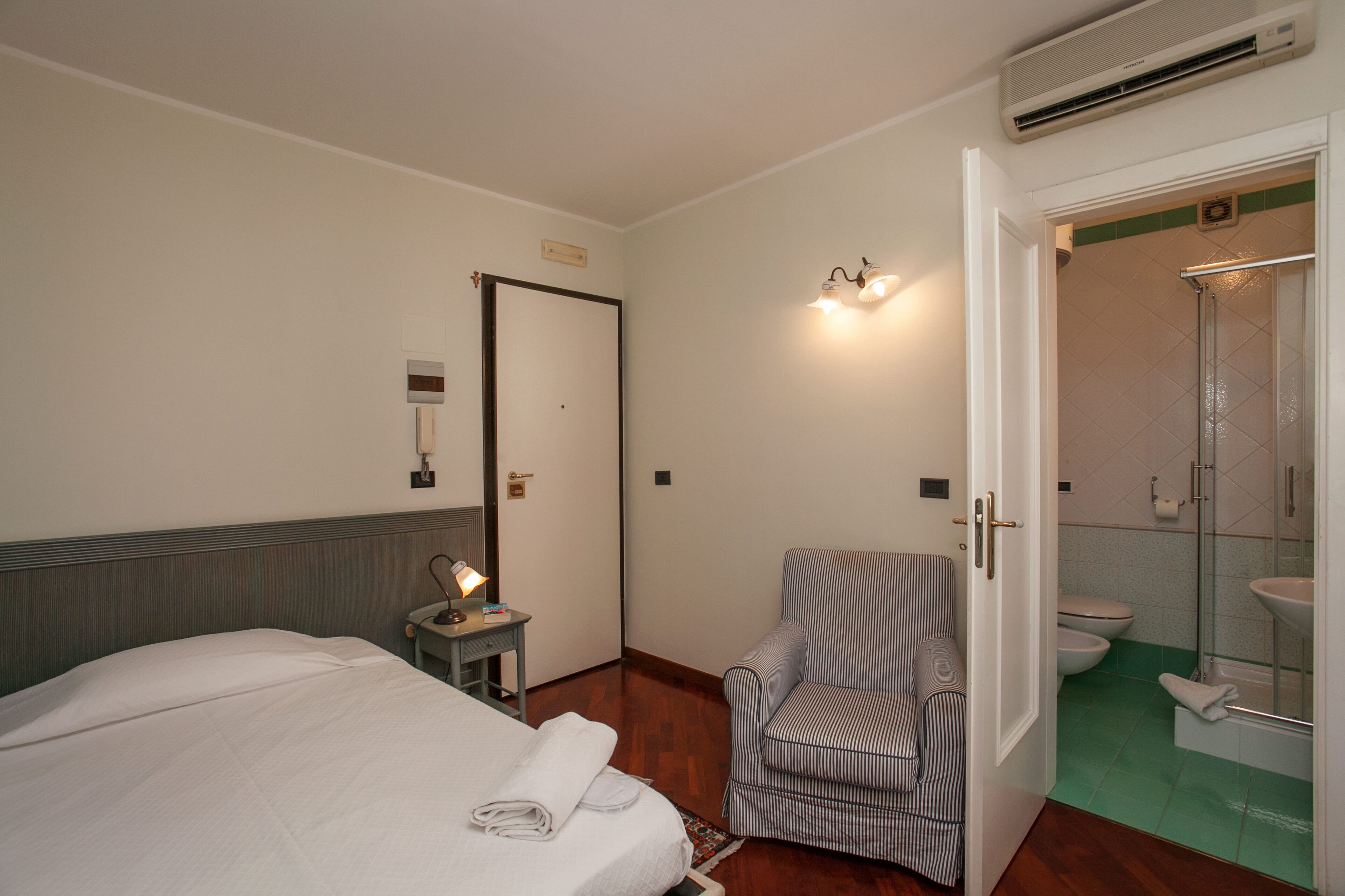 Et hotelværelse med seng, stol, lille bord med lampe, badeværelse med bruser og toilet.