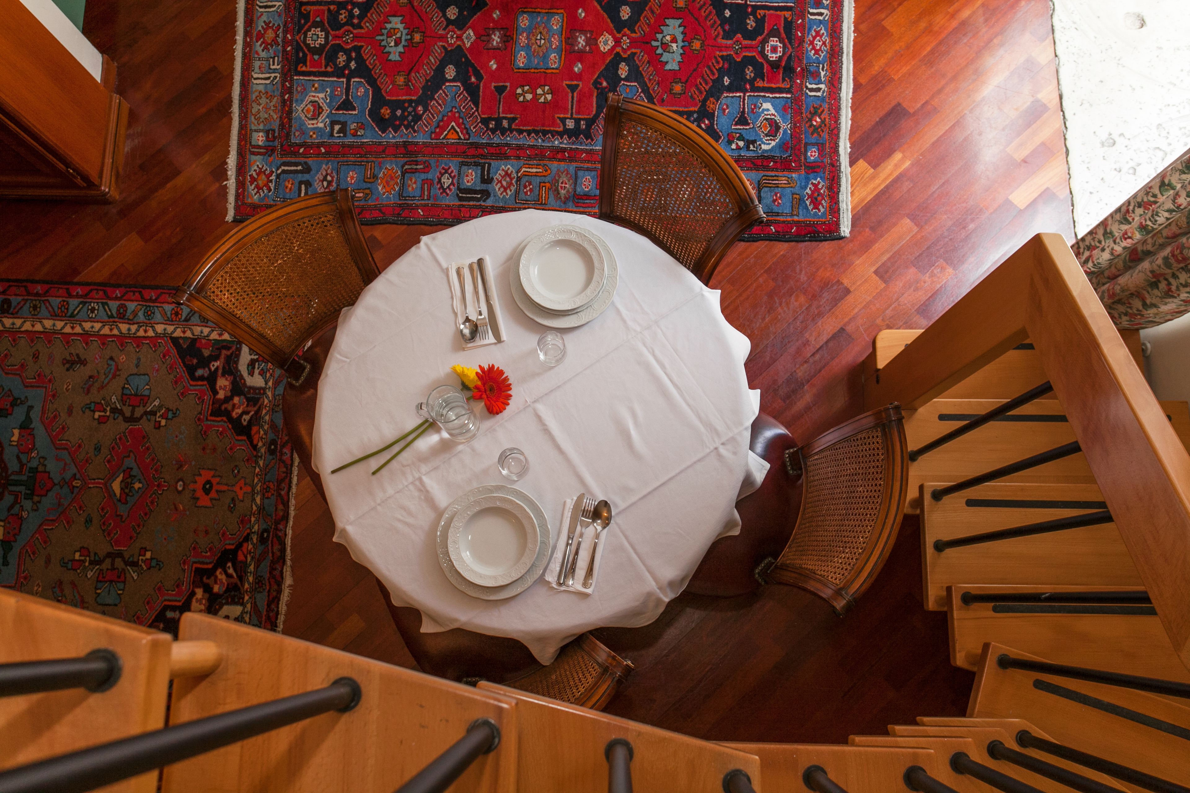Una mesa redonda de comedor con mantel blanco, dispuesta con platos, cubiertos y un jarrón con flores. La mesa está rodeada de sillas de madera.