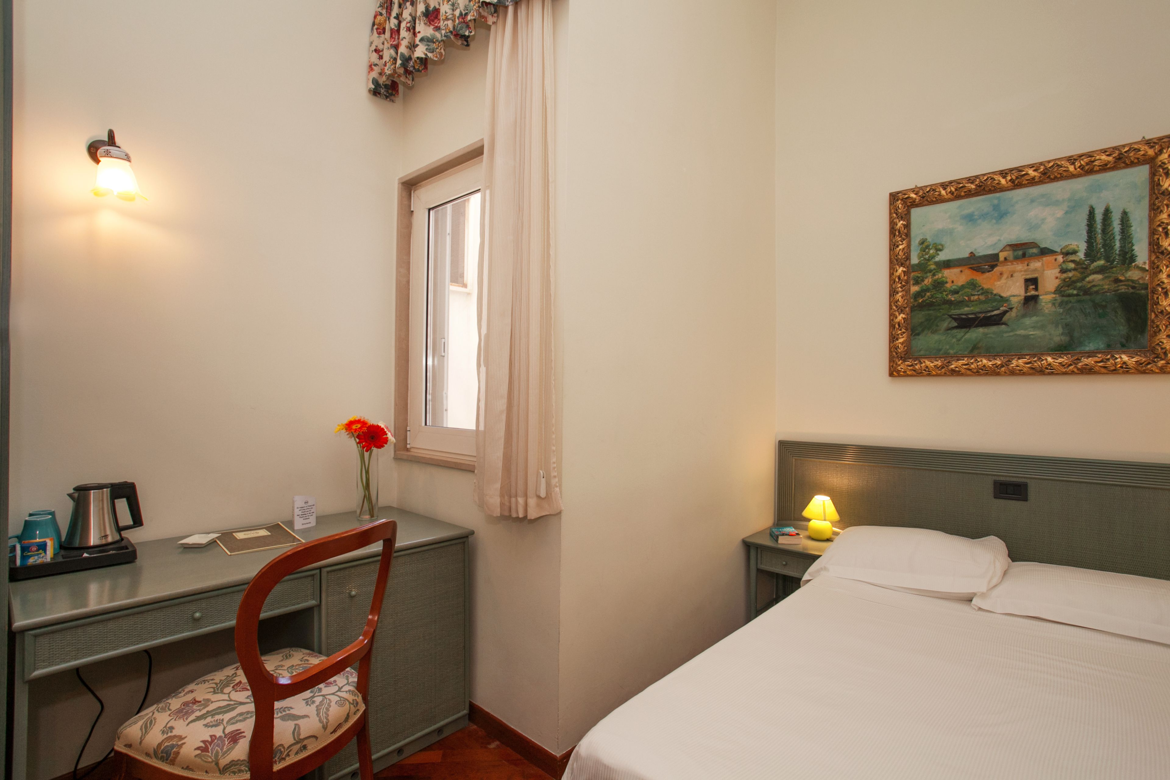 A hotel room with a bed, a desk with a chair, a painting on the wall, and a window with curtains.
