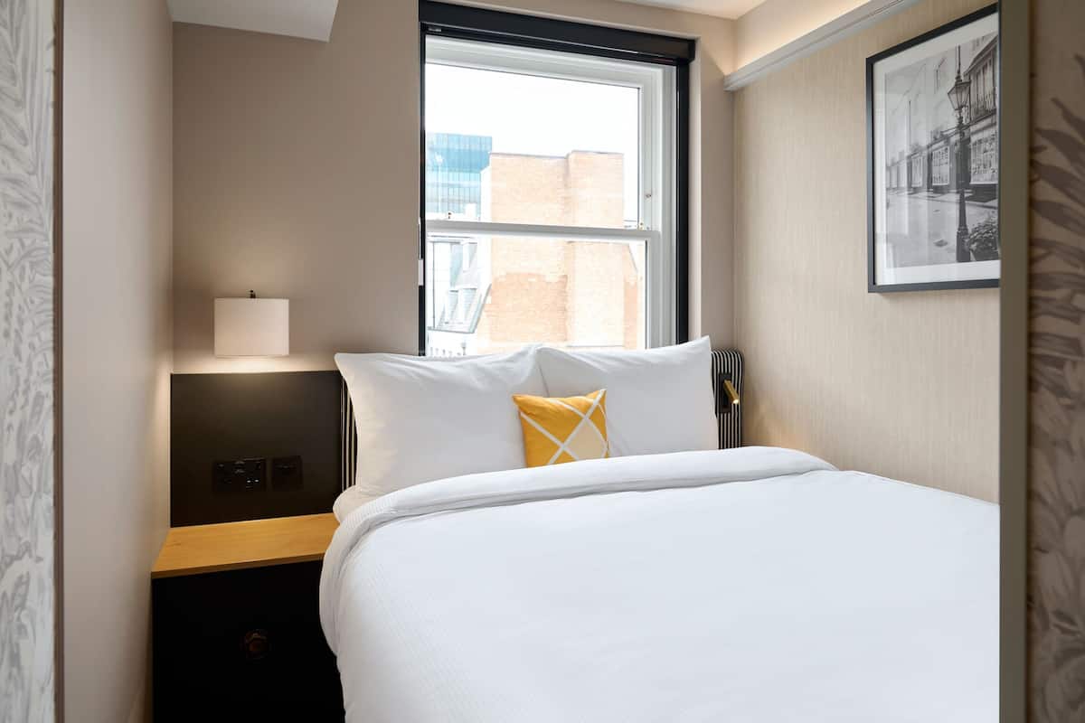 Une chambre d’hôtel avec un lit, une table de chevet, une lampe et une fenêtre donnant sur des immeubles.