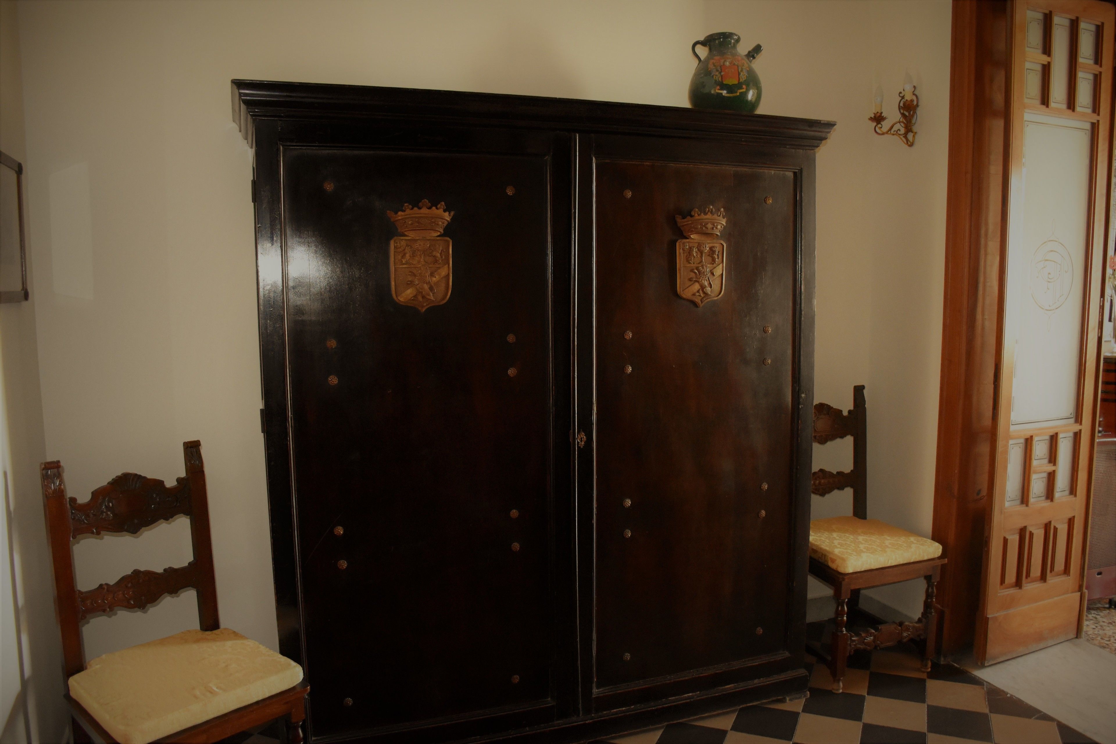 A room with a large wooden wardrobe featuring two coats of arms, a wooden chair with a cushion, and a checkered floor.