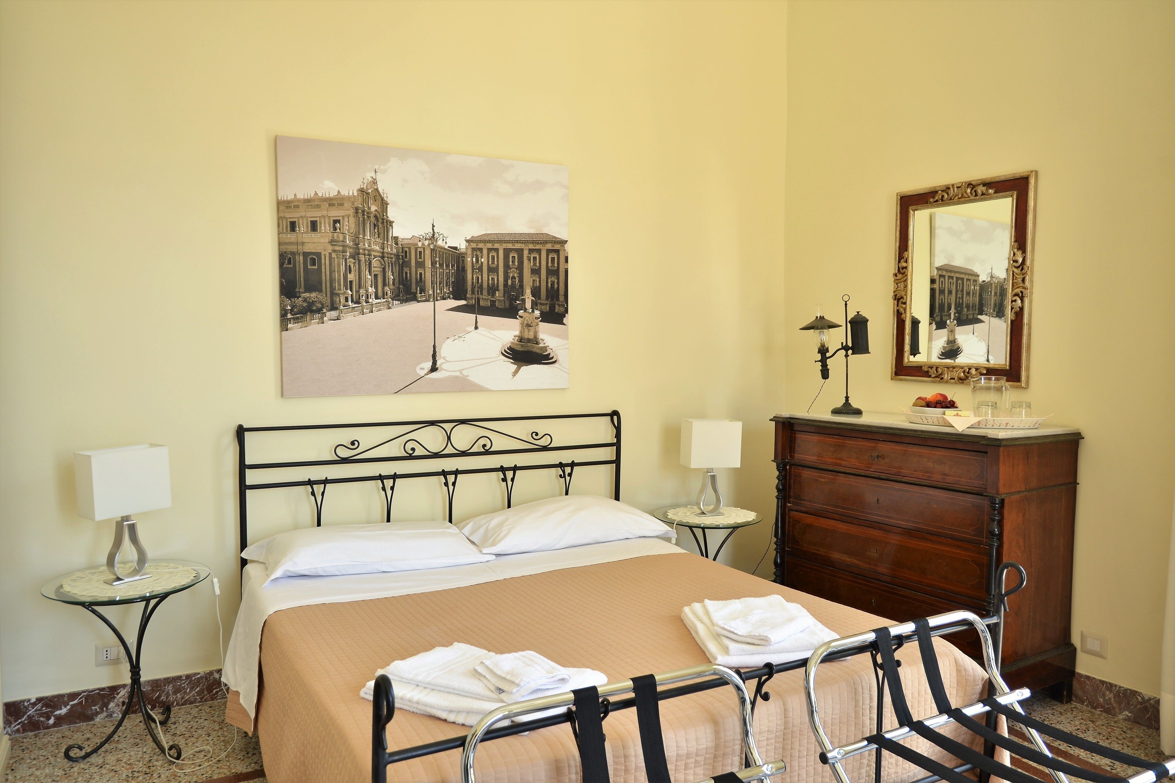 A hotel room with a bed, two bedside tables, a dresser, a mirror, and a painting of a cityscape.