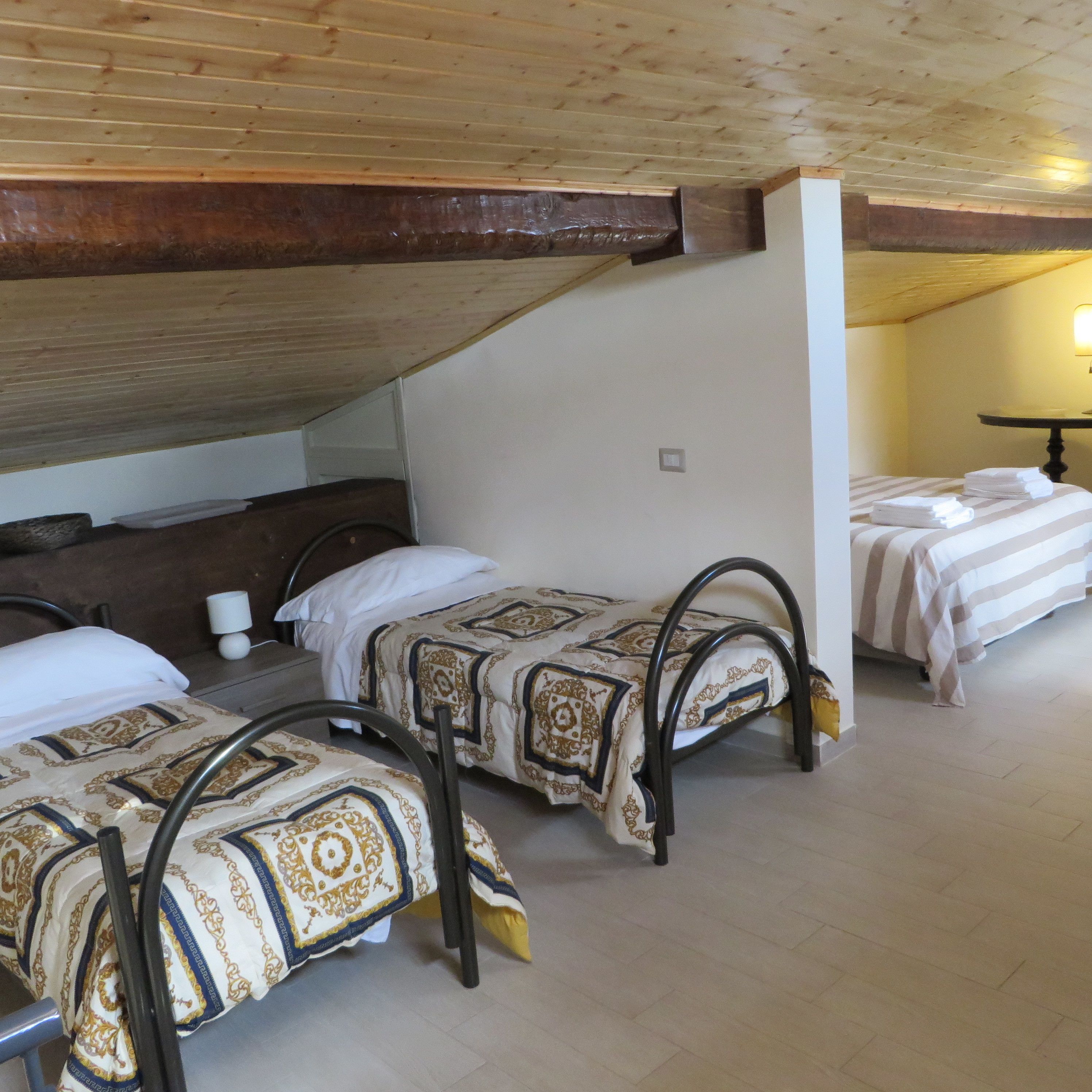 A dormitory room with three beds, wooden beams on the ceiling, and a small table with a lamp.
