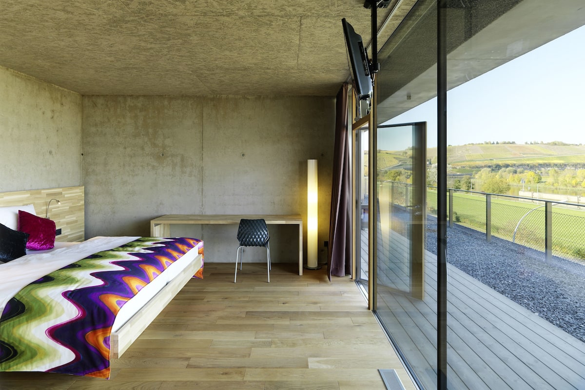 A bedroom with a bed, a desk, and a large window offering a view of the countryside.