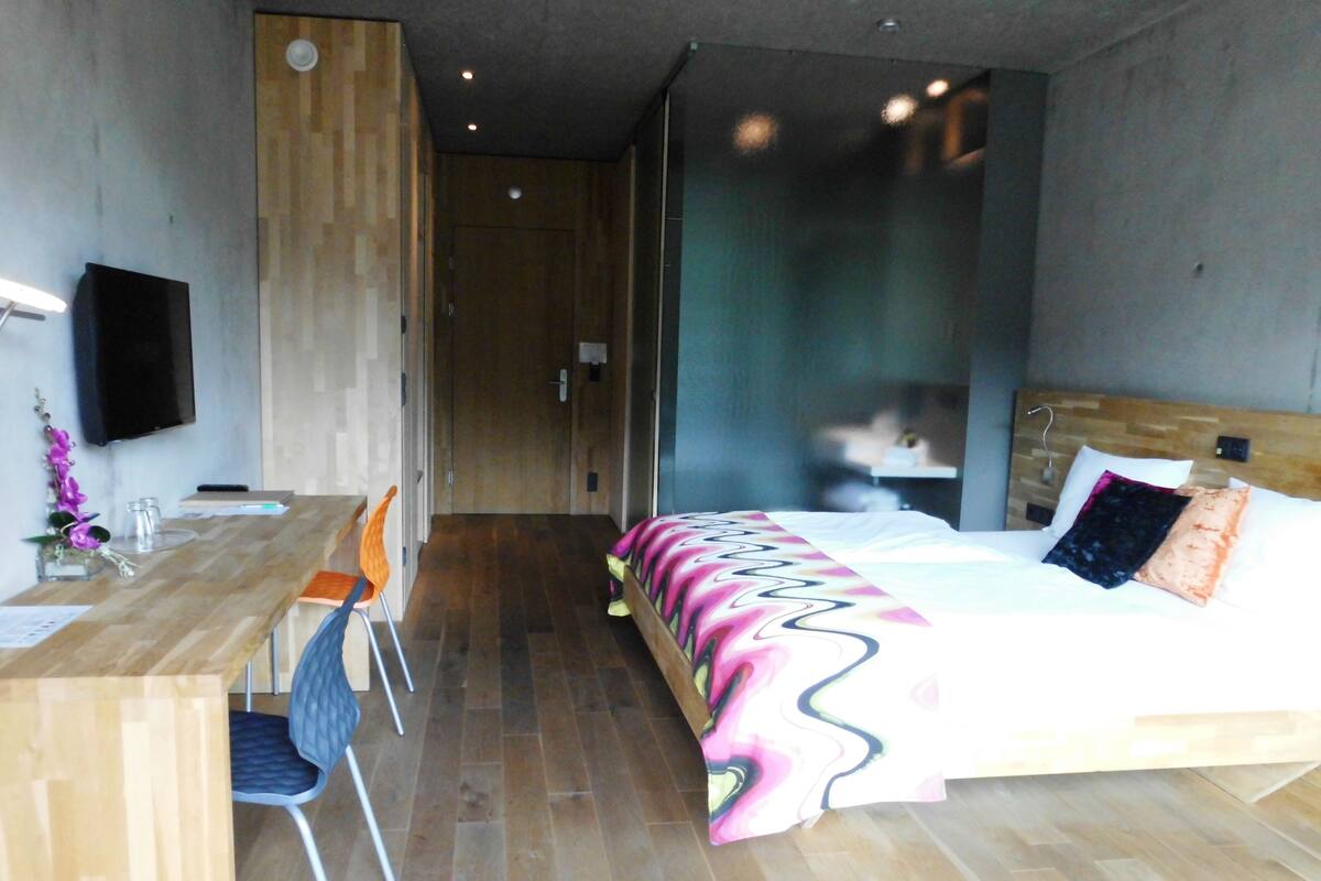 A modern hotel room with a wooden floor, a desk with a chair, a bed with a colorful blanket, and a TV mounted on the wall.