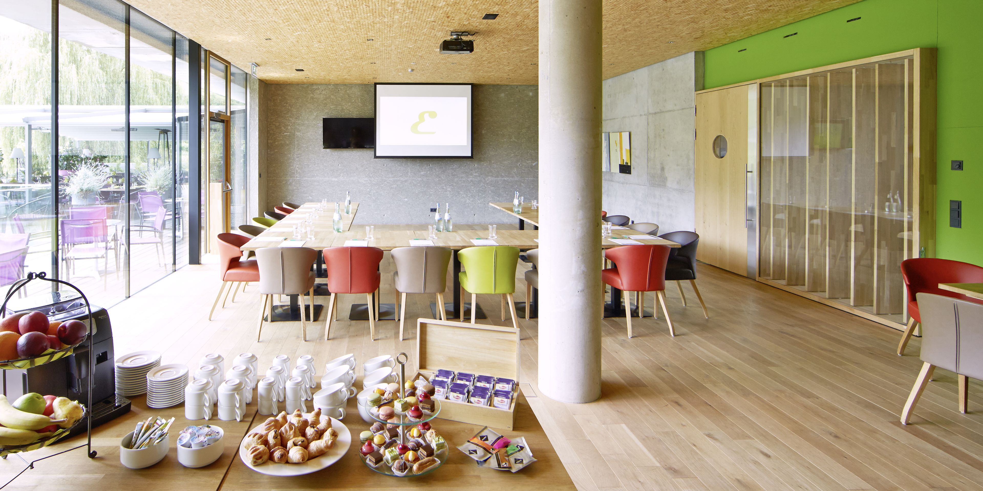 A modern conference room with a long table, colorful chairs, and a variety of food and drink items on the table.