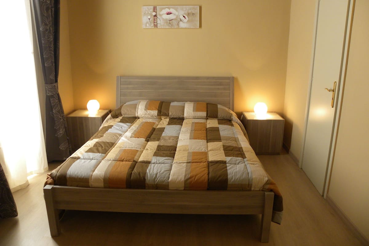 A neatly made bed with a quilt, two bedside lamps, a headboard, and a wall-mounted artwork.