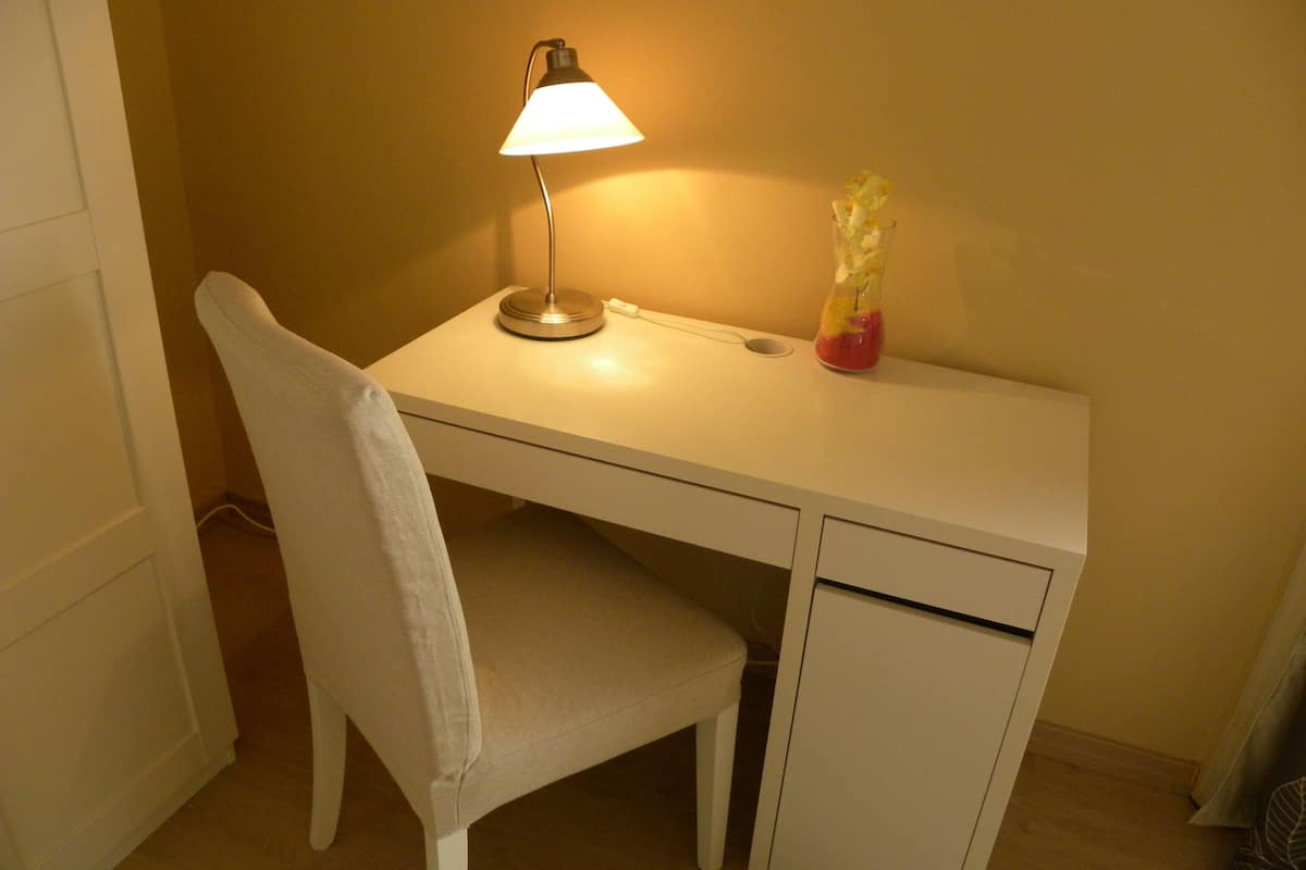 A small, white desk with a lamp and a vase of flowers.