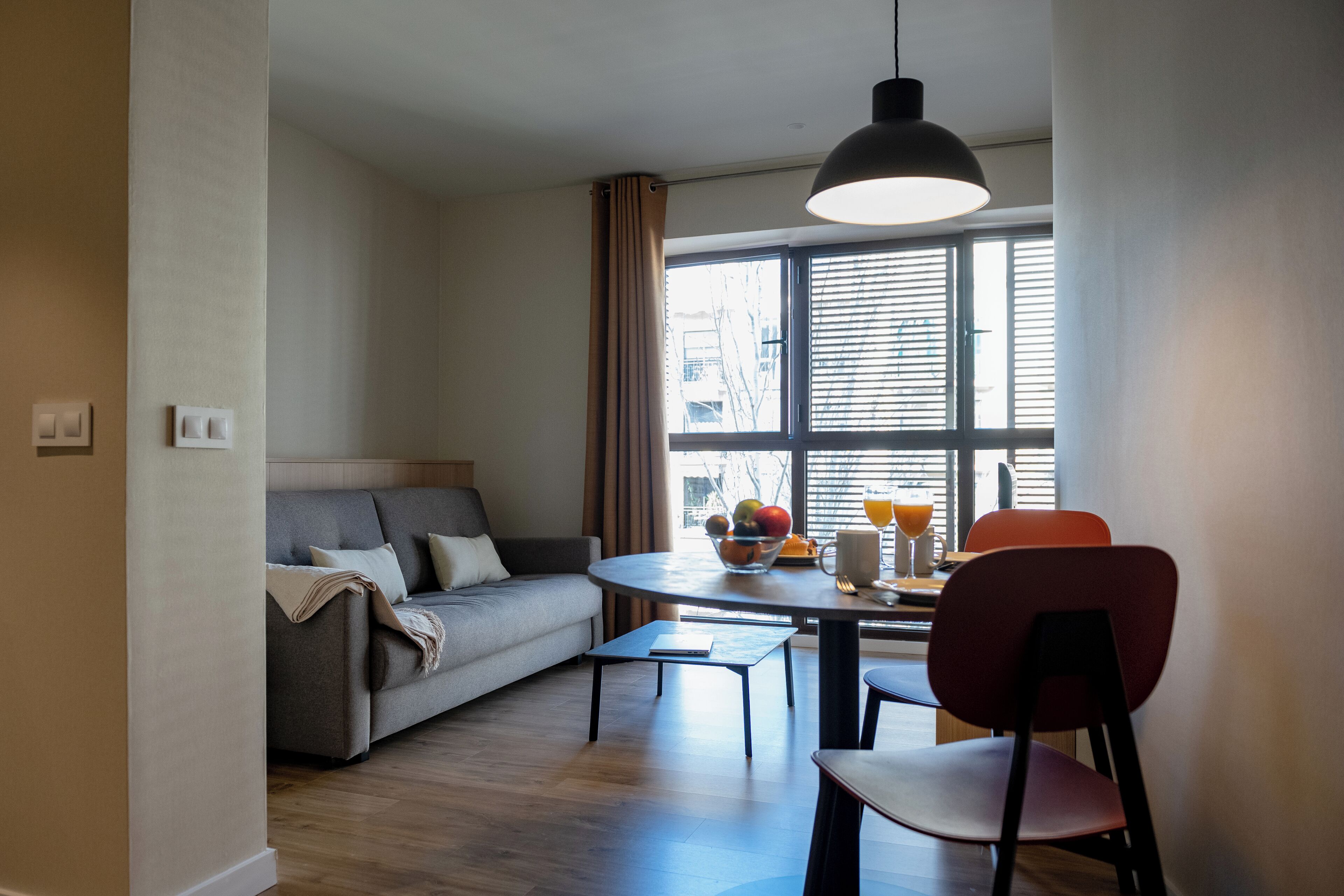 A modern living room with a sofa, dining table, and chairs. There is a window with blinds, a hanging light fixture, and a bowl of fruit on the table.