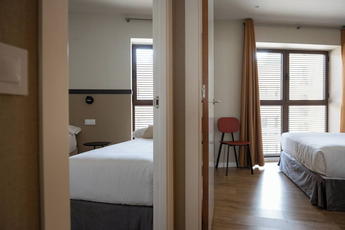 A hotel room with two beds, a small table, a chair, and a window with blinds.