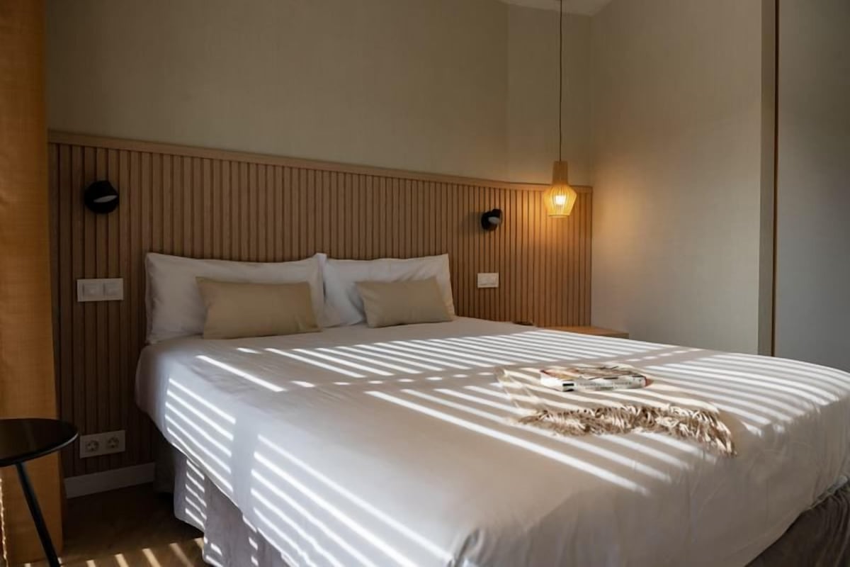 A neatly made bed with a white comforter and pillows, a bedside table with a lamp, and a wall-mounted light fixture.