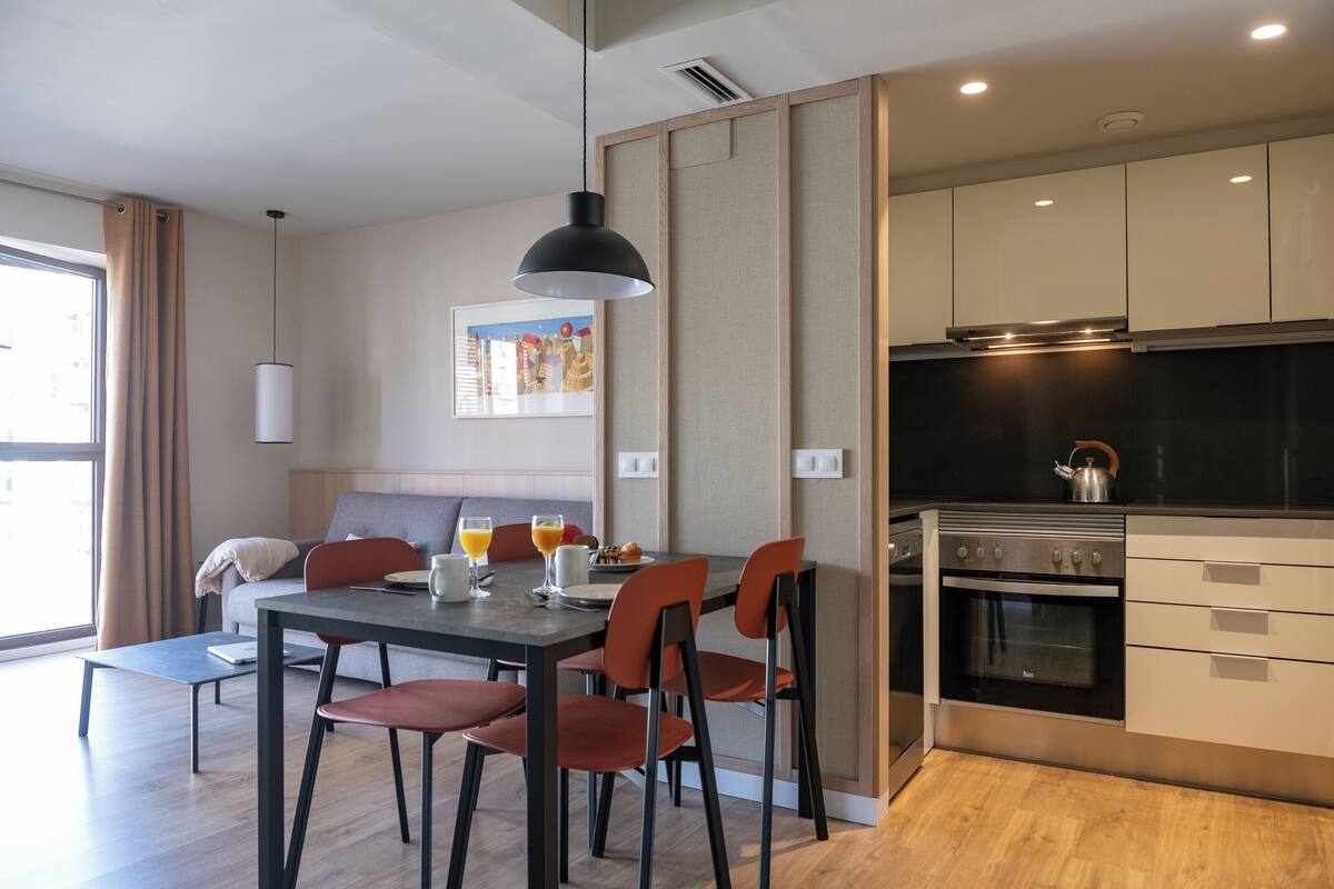 A modern kitchen with a dining area, wooden flooring, and a large window with curtains.