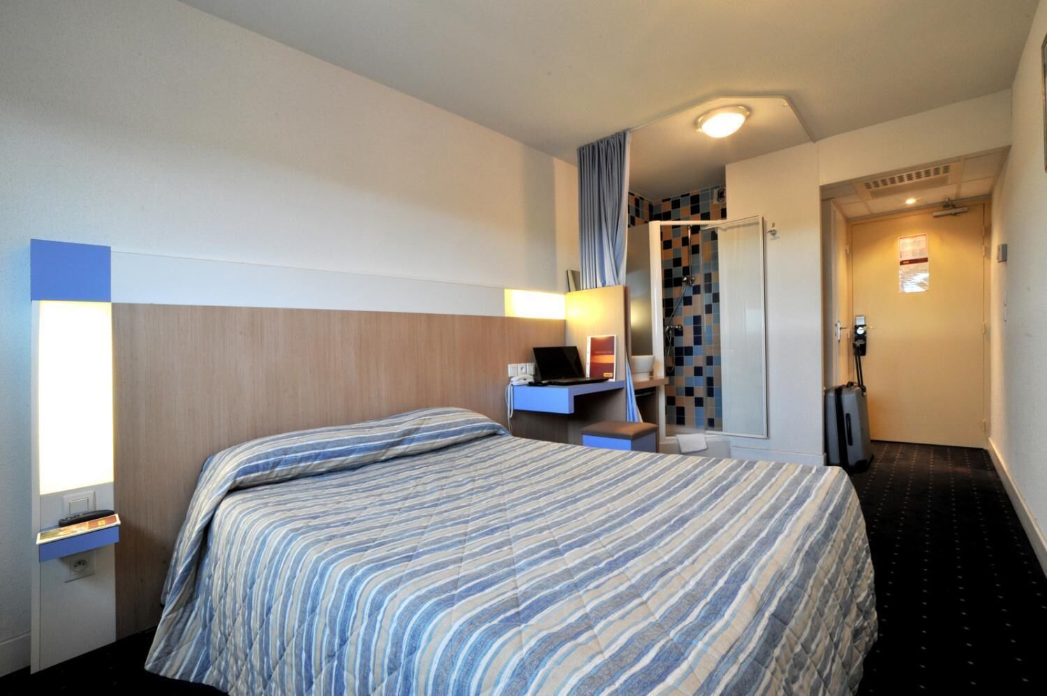 A hotel room with a bed, a desk with a laptop, a shower area, and a door with a keycard reader.