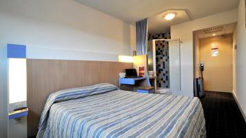 A hotel room with a bed, a desk with a laptop, a shower area, and a door with a keycard reader.