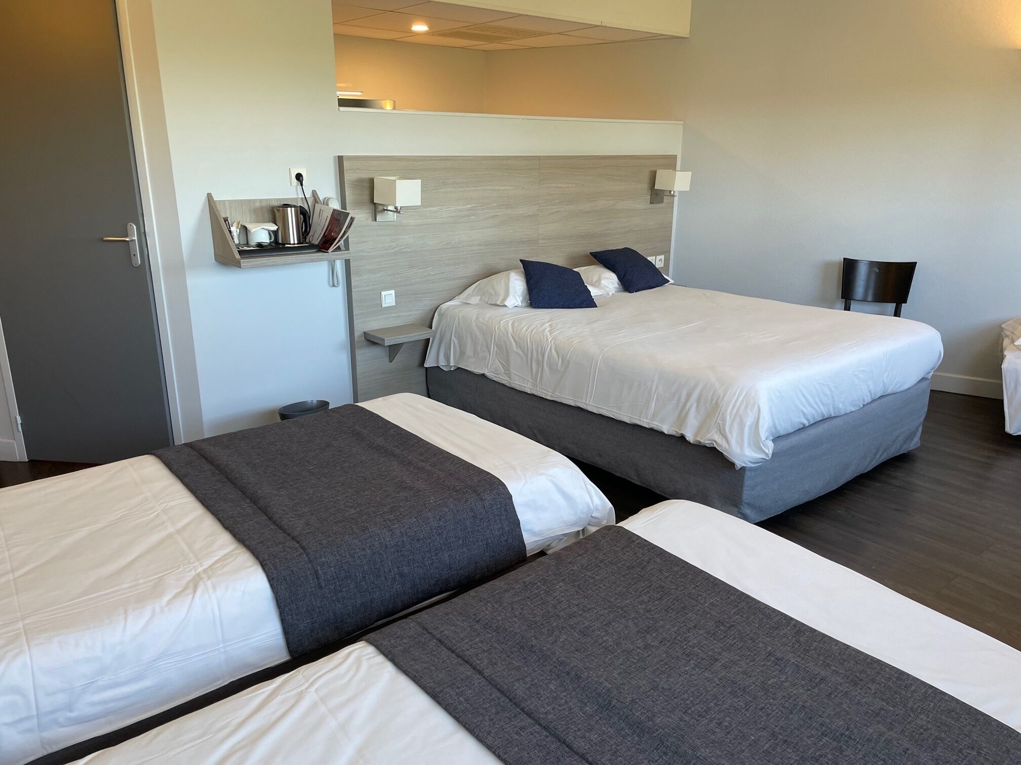 Habitación de hotel con dos camas, un escritorio pequeño con lámpara y estante en la pared con artículos de aseo.