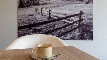 A table with a cup of coffee and a plate, a chair, and a black and white photograph of a rural landscape on the wall.