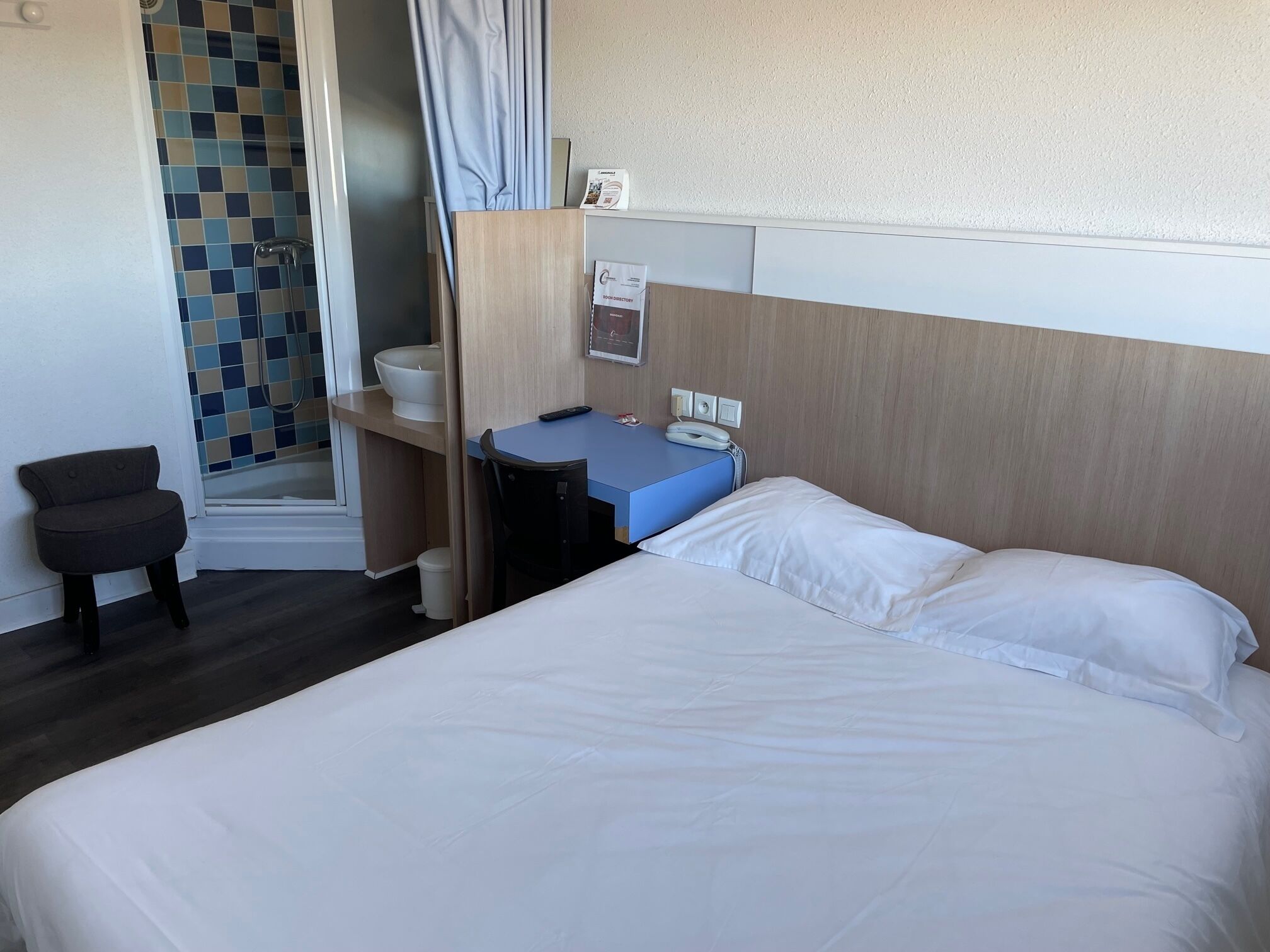 A hotel room with a bed, a desk, a chair, a bathroom with a shower, and a small stool.