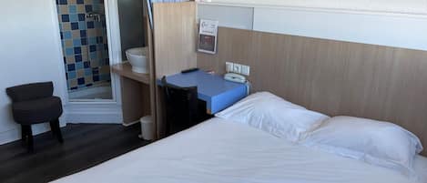 A hotel room with a bed, a desk, a chair, a bathroom with a shower, and a small stool.